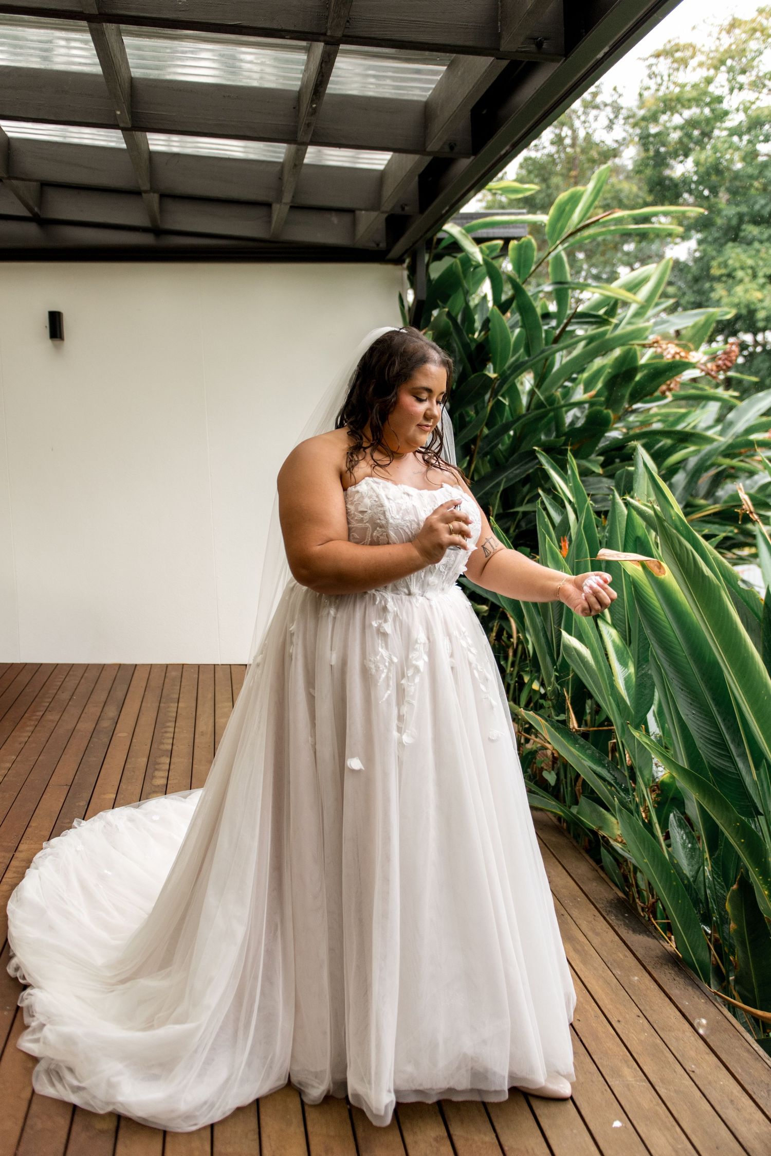 Bride in a strapless lace ball gown with long train standing on a deck surrounded by tropical plants.