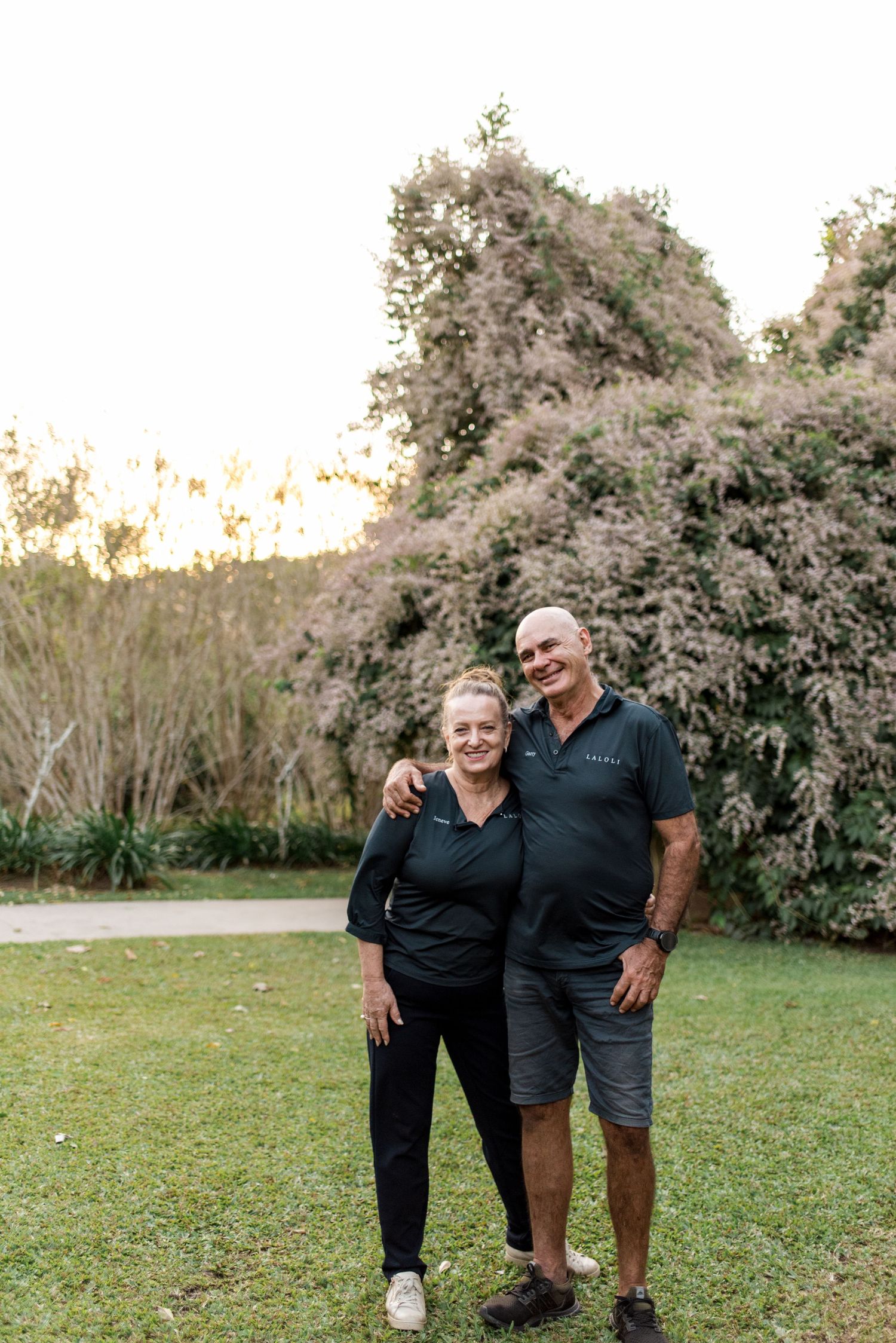 Happy mature couple standing together on green lawn in front of lush garden trees.