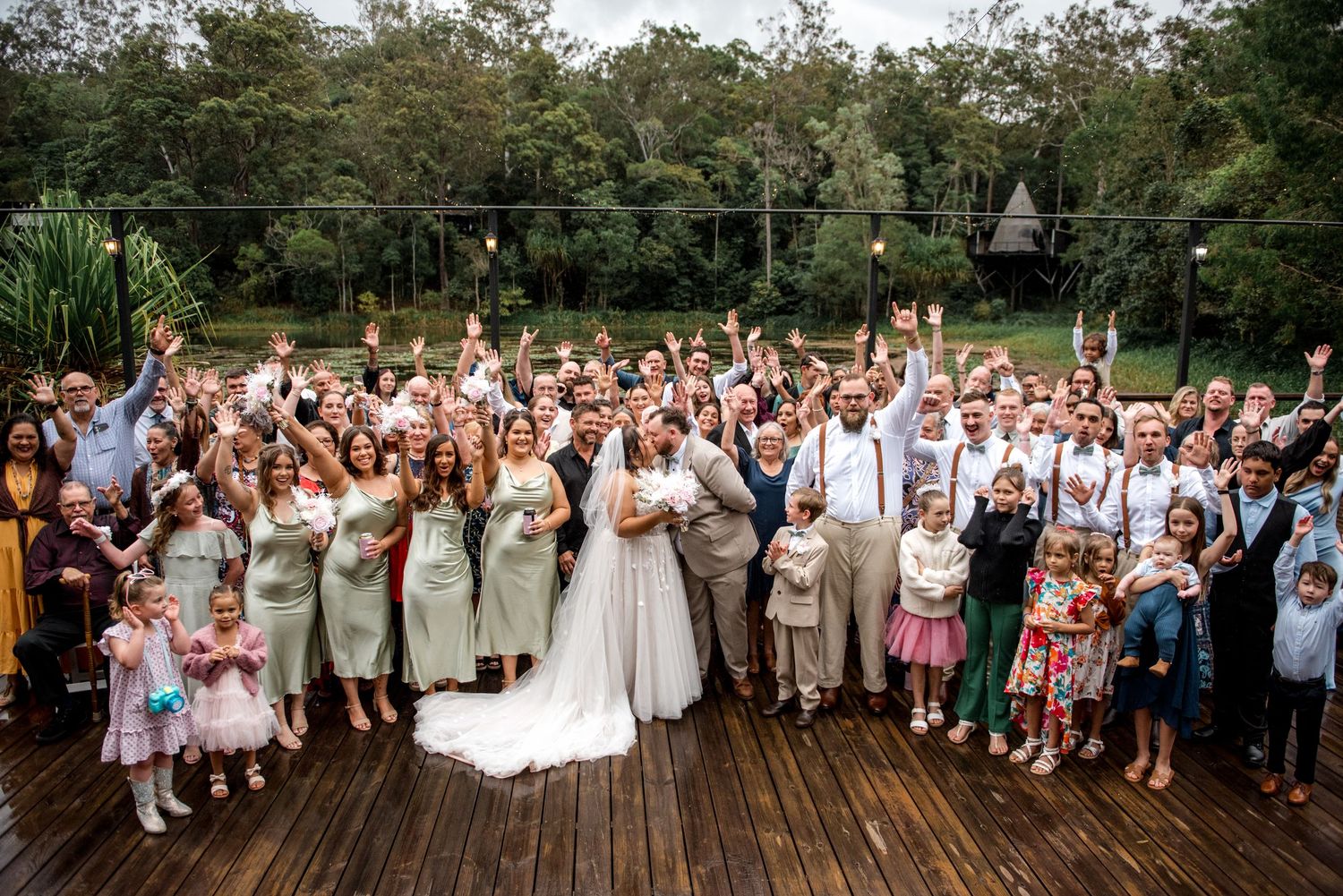 Large wedding group photo with bride and groom surrounded by smiling guests on a wooden deck with lush greenery.