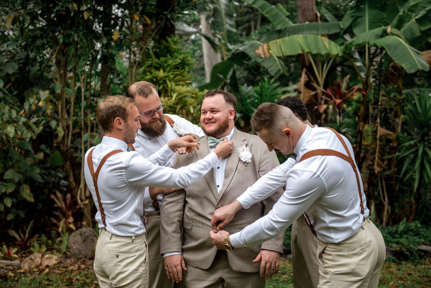 Groomsmen in white shirts and suspenders help groom in tan suit with his bow tie outdoors.