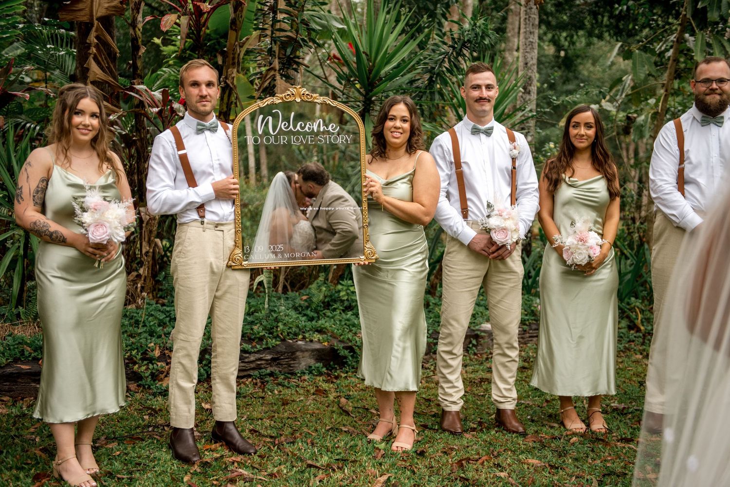 Wedding party poses with sage green bridesmaid dresses and suspenders beside a welcome mirror sign.