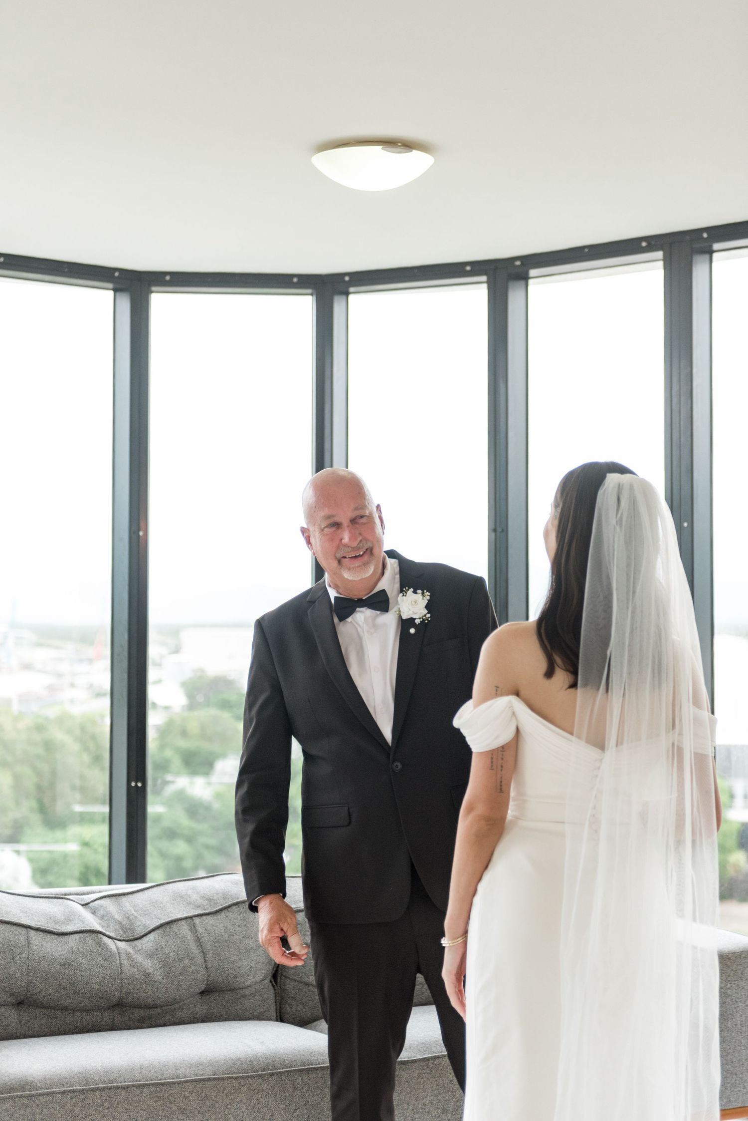 Groom in black tuxedo smiles at bride in white off-shoulder gown during first look in a bright windowed room.