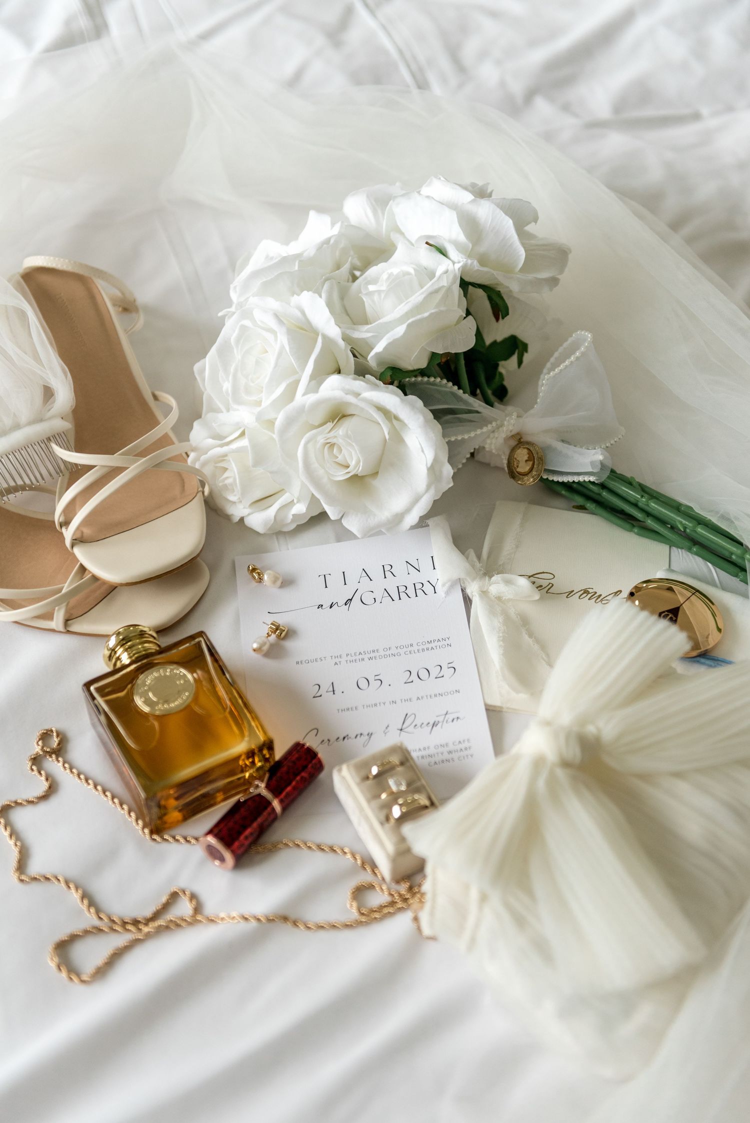 Bridal flat lay with white roses bouquet, nude strappy heels, gold perfume, wedding invitation, and accessories on white linen.