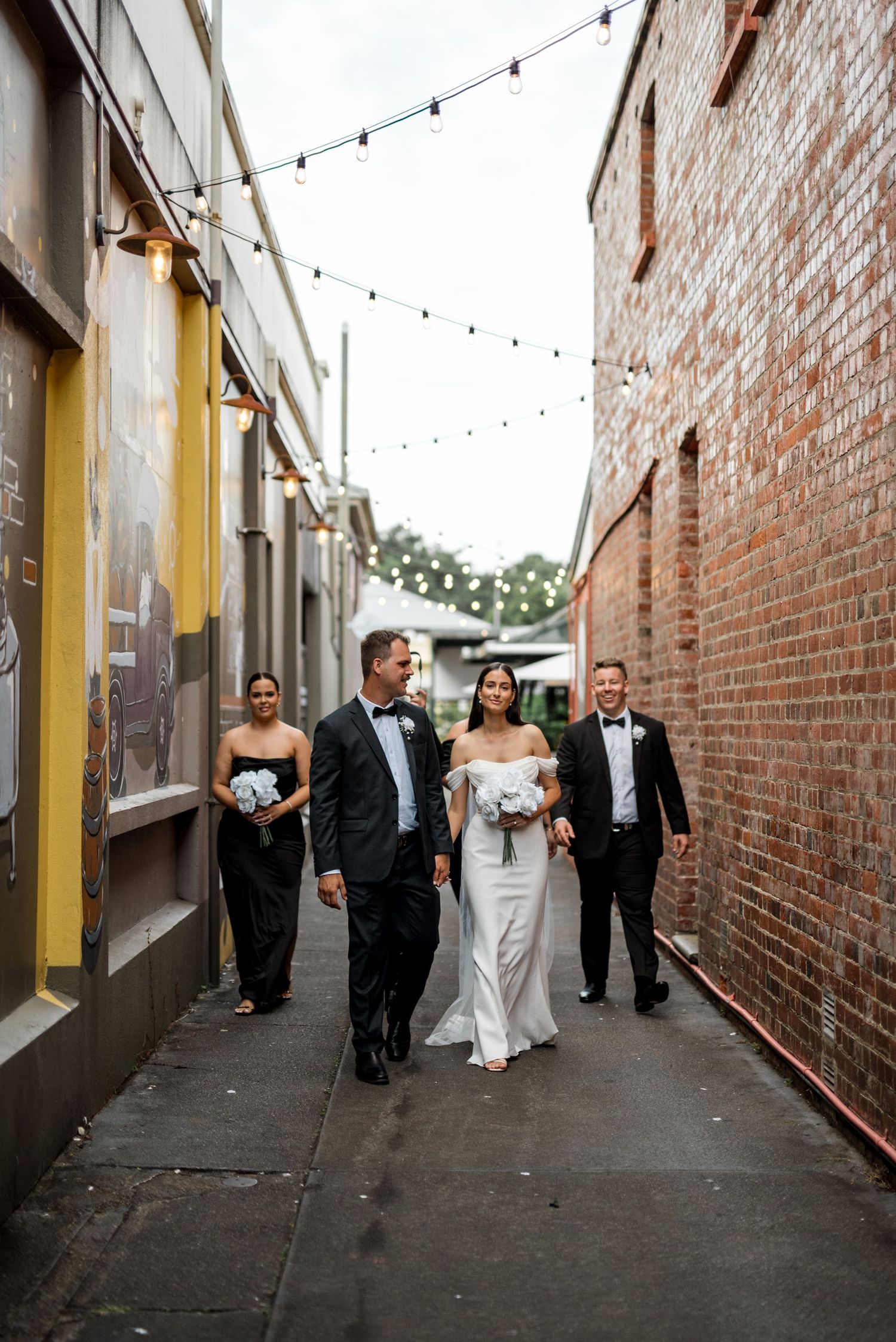 Bride and groom with wedding party walking through a festoon-lit alleyway.
