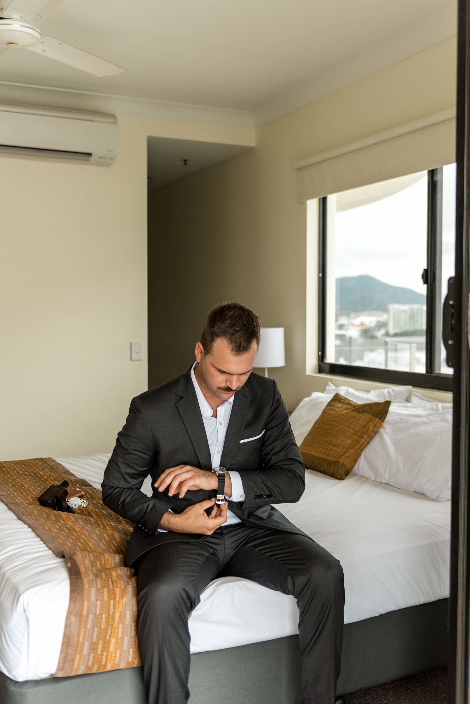 Groom in dark suit fastens his watch while seated on a hotel bed with a scenic mountain view window behind him.