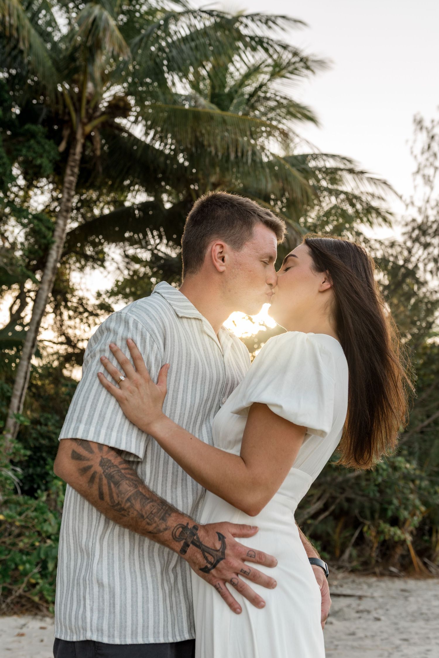 Tattooed couple sharing a passionate kiss on a tropical beach during a warm golden sunset.