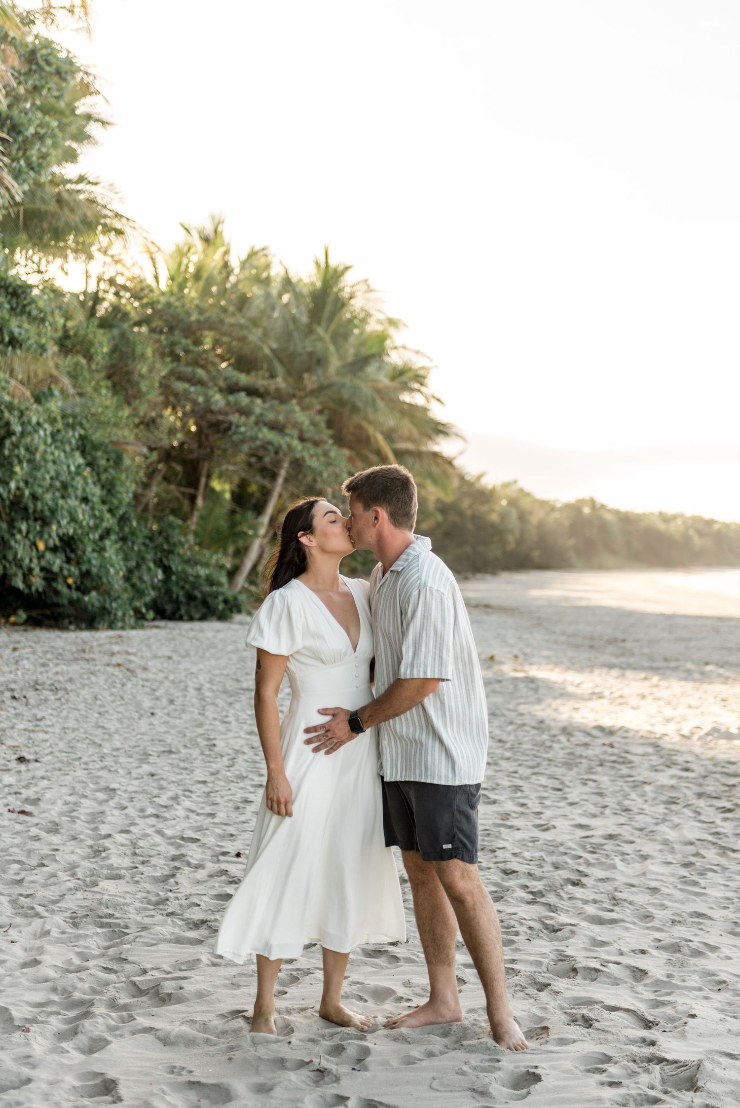 Couple sharing a kiss on a sandy tropical beach at sunset with lush green trees behind them.