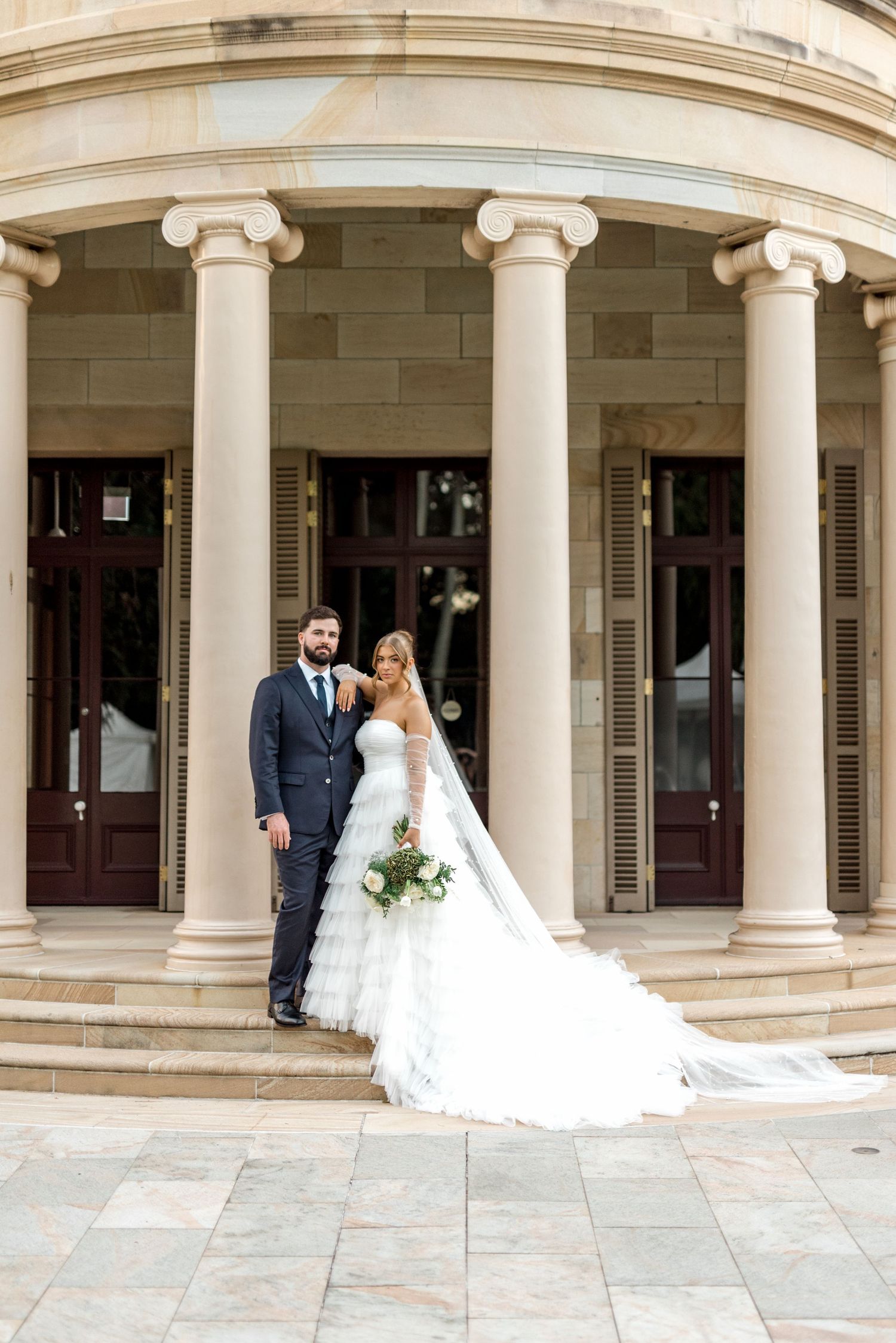 Bride and groom posing on steps of elegant colonial building with ionic columns.