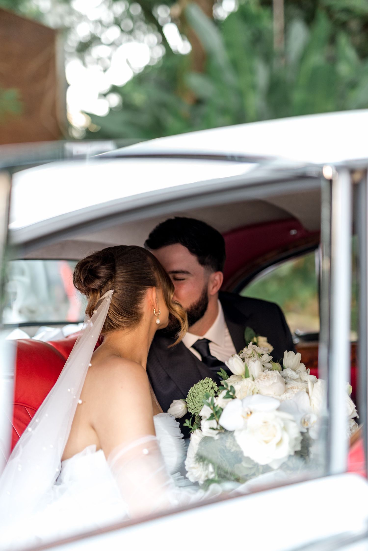 Bride and groom share a kiss inside vintage car with red interior, bride holding white floral bouquet.