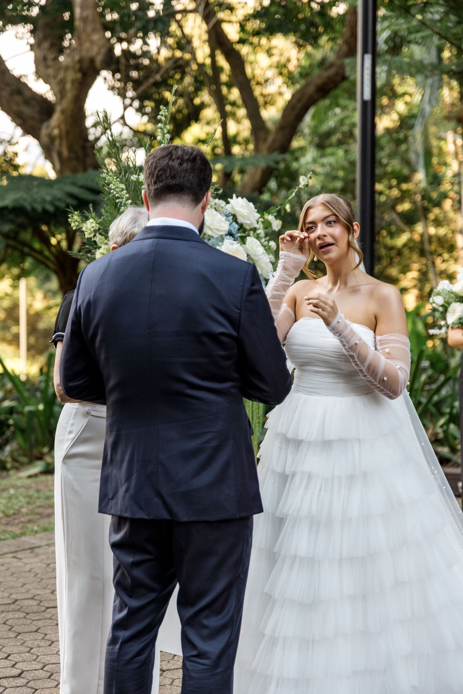 Bride wiping tears during outdoor wedding ceremony in white tiered gown with sheer gloves.