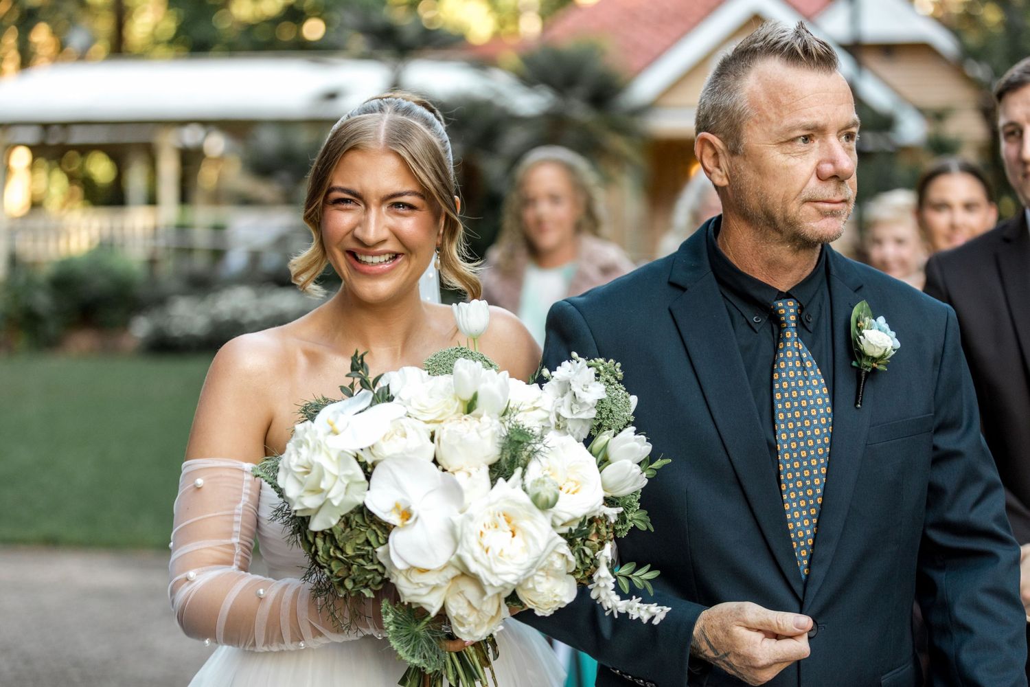 Smiling bride in strapless tulle gown holds white rose bouquet while walking with escort.