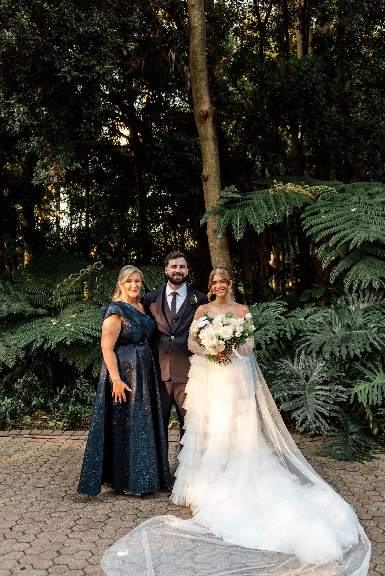 Bride in white gown with veil and groom in brown suit pose with a woman in navy dress in a lush garden.