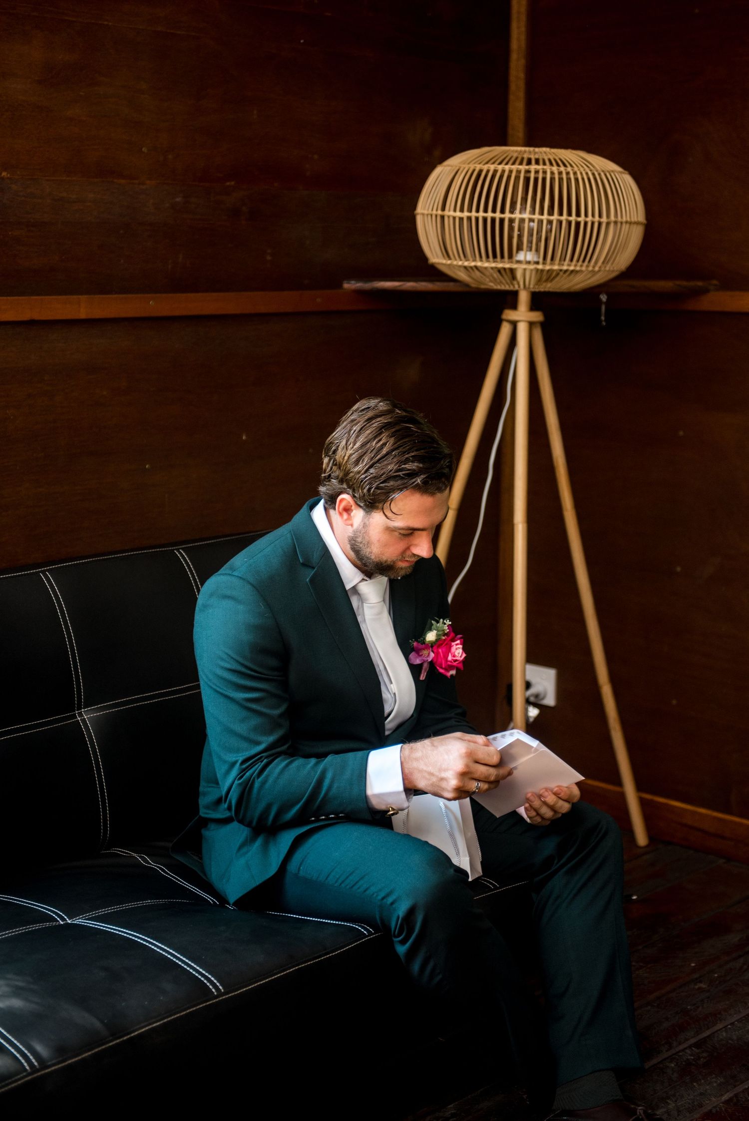 Groom in teal suit reading letter on leather couch before wedding ceremony.
