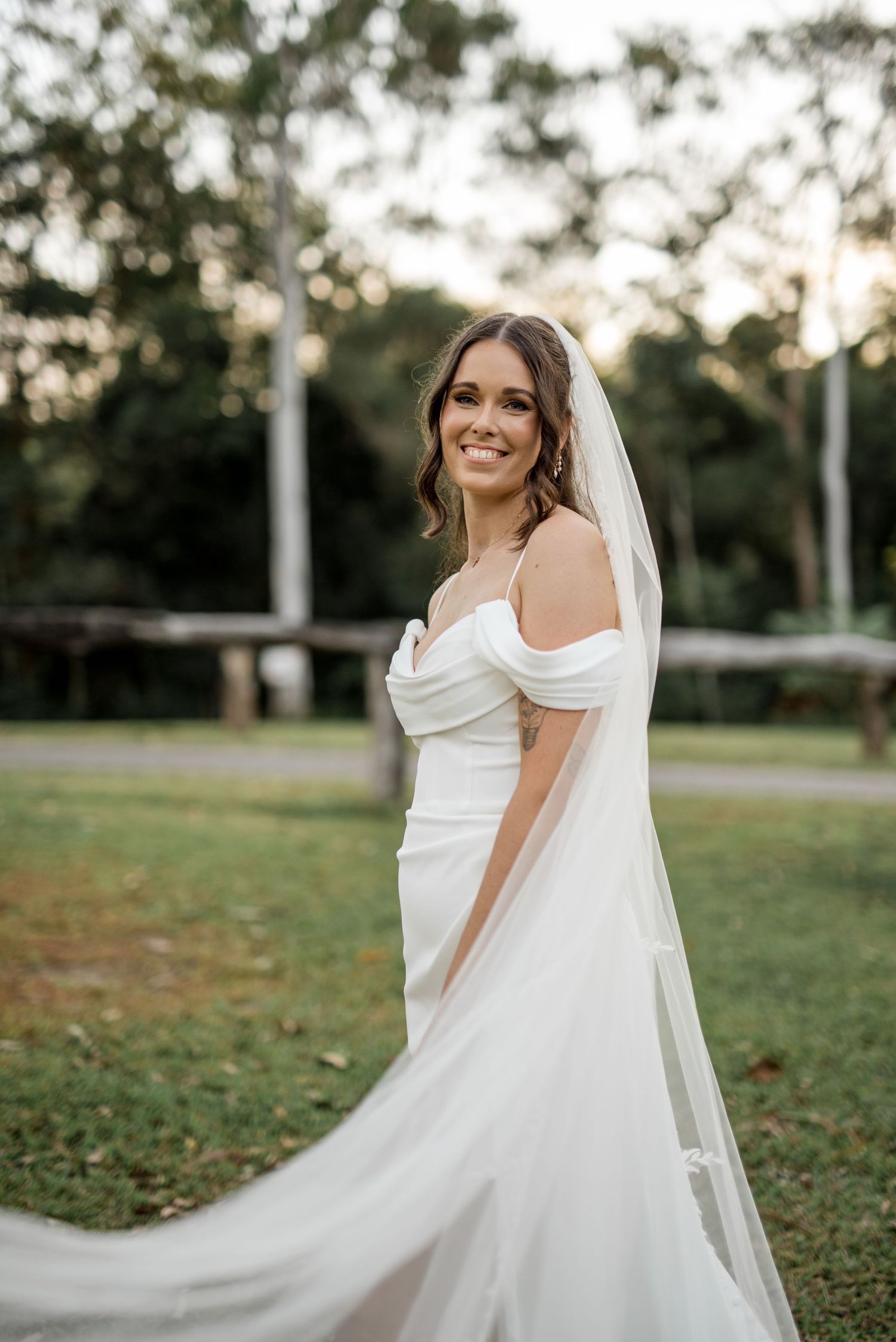 Radiant bride in off-shoulder wedding gown and veil smiling in a lush garden setting.
