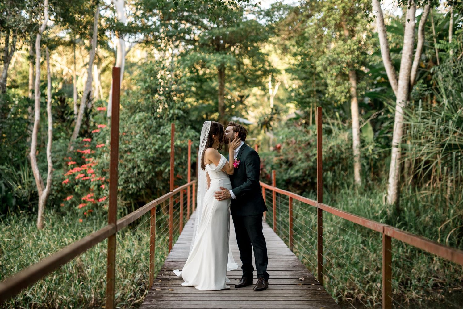 Bride and groom share a kiss on a wooden bridge surrounded by tropical forest greenery.