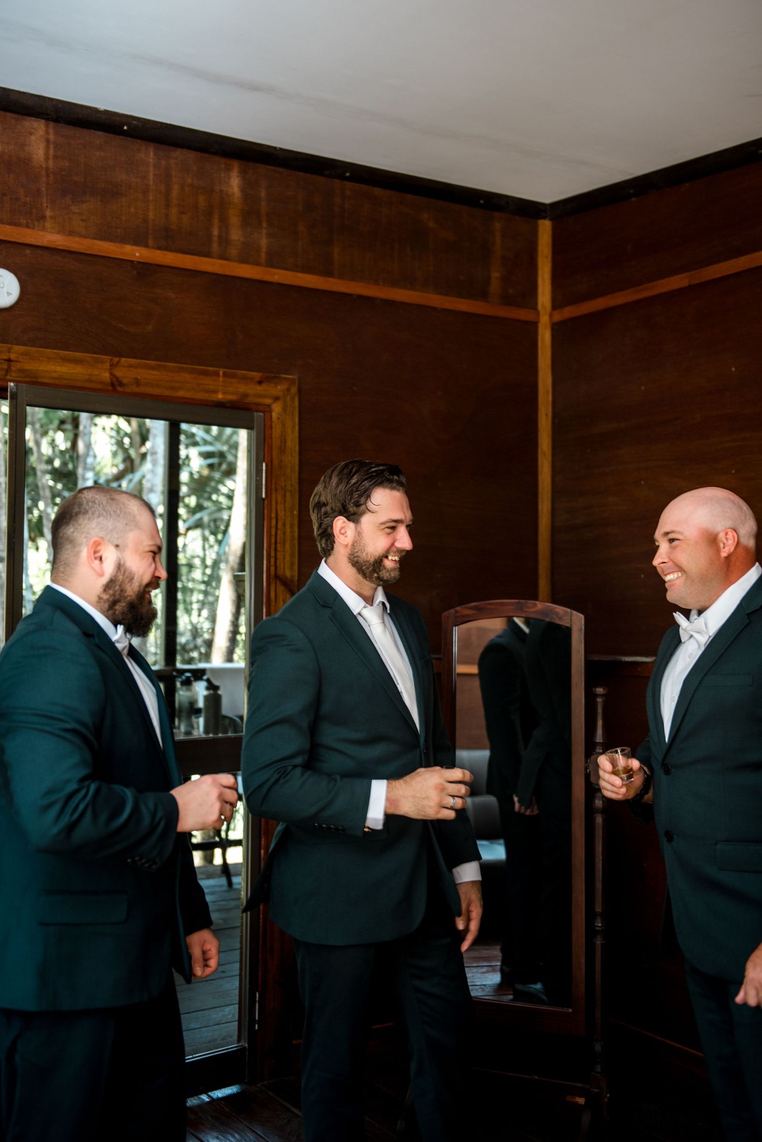 Three groomsmen in dark suits laughing and chatting together while getting ready in a wood-paneled room.