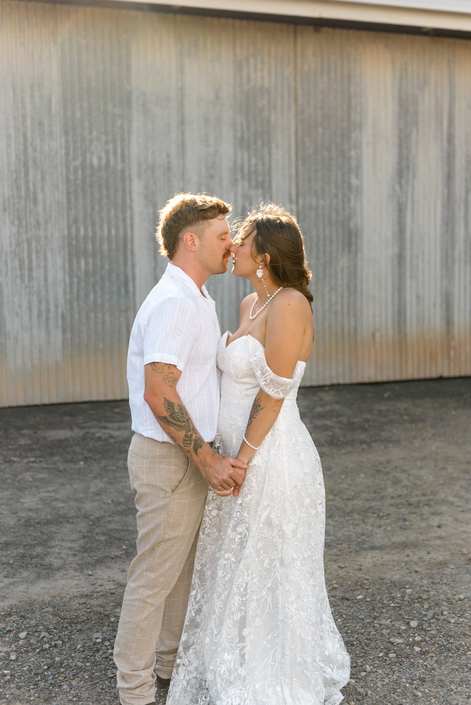 Bride and groom lean in for a kiss while holding hands, bathed in warm sunlight against a metal wall.