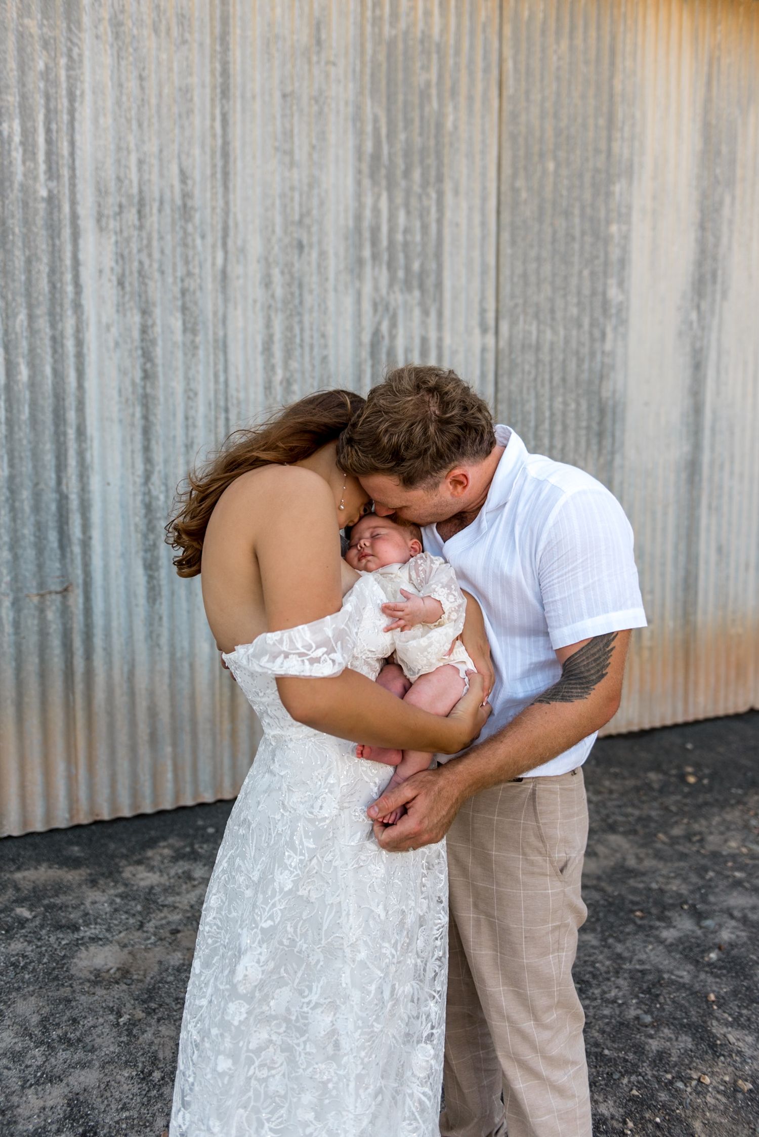 Bride and groom in wedding attire sharing a kiss over their newborn baby against metal wall.