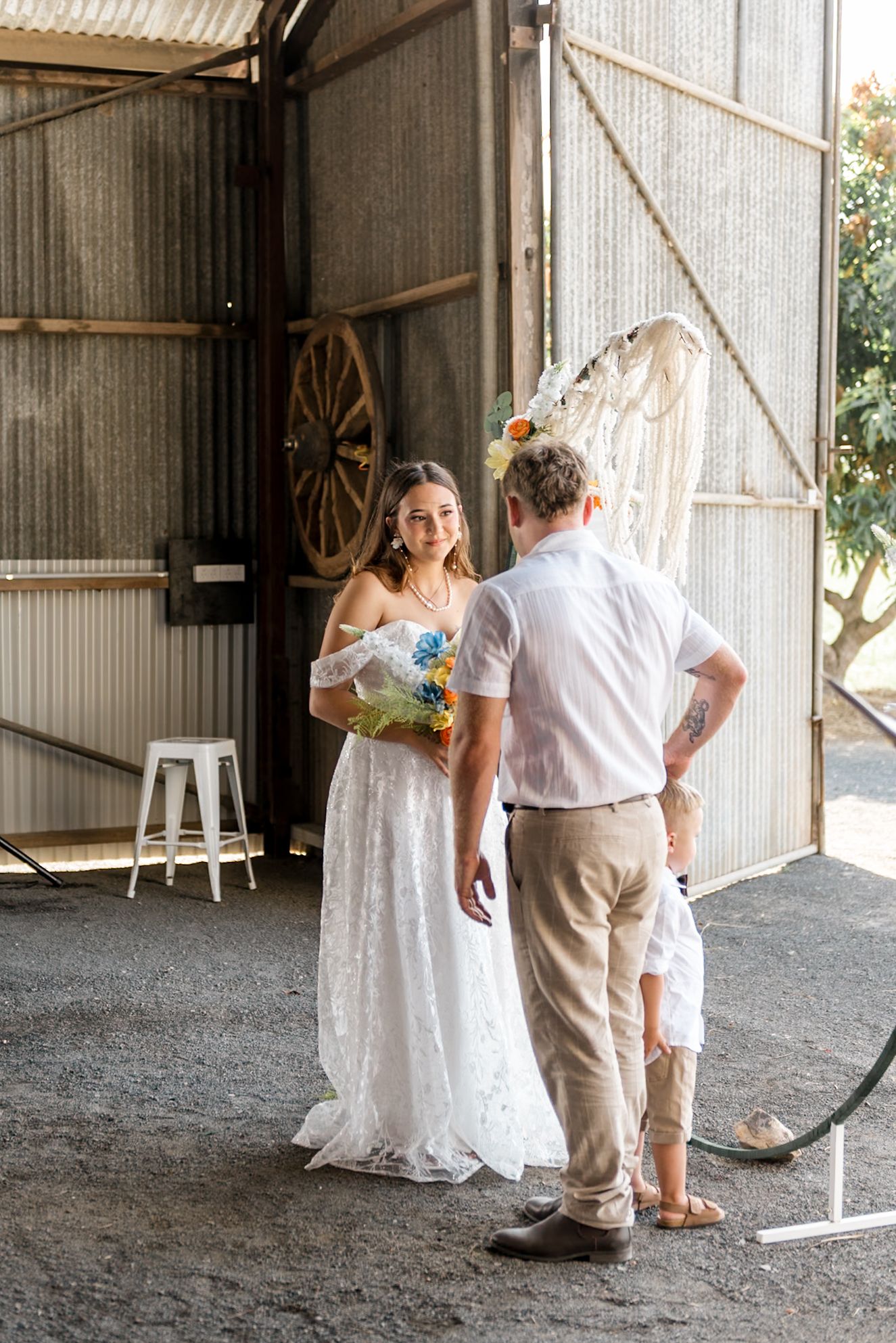 Smiling bride in off-shoulder lace gown holds colorful bouquet facing groom in rustic barn.