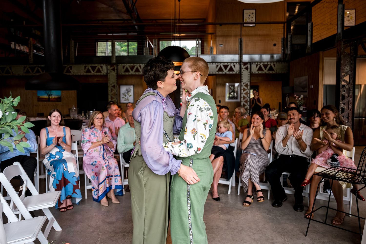 Two brides embrace and kiss at wedding ceremony while smiling guests applaud in white chairs behind them.