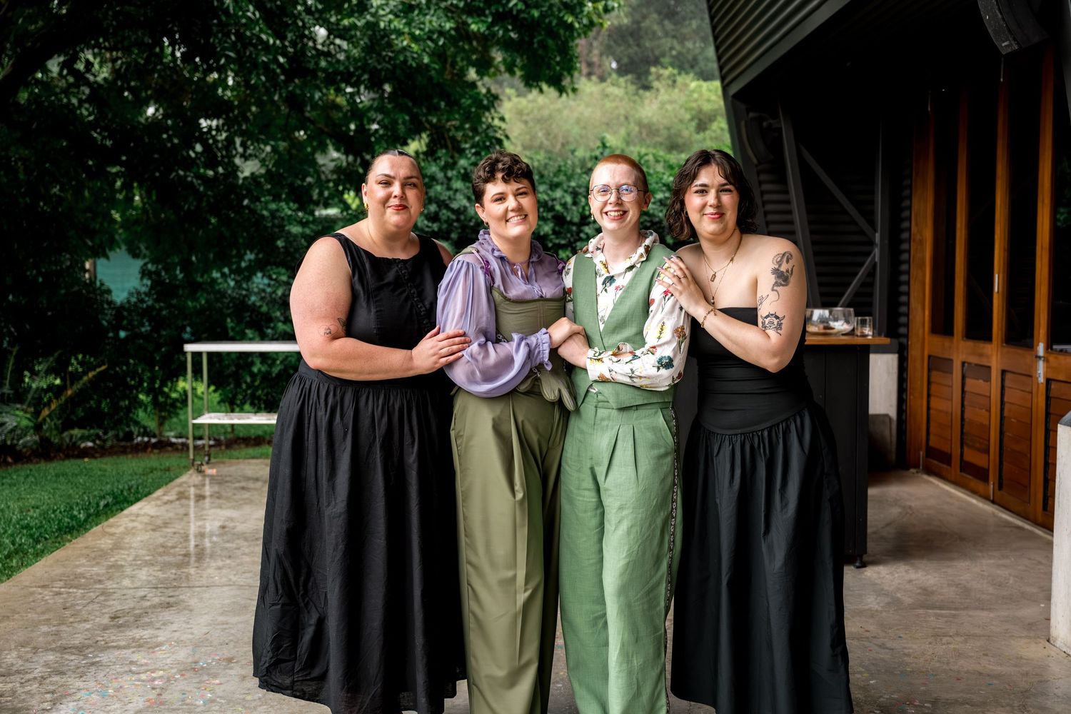 Four women smiling together outdoors at a wedding celebration surrounded by lush greenery.