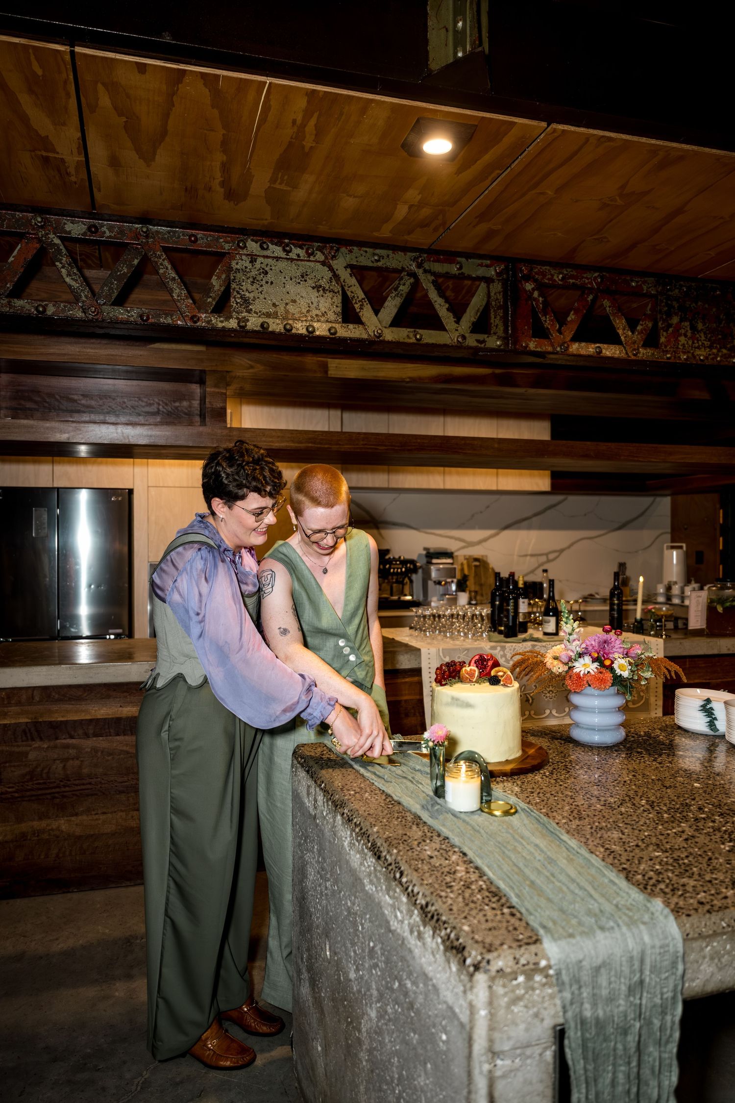 Two people in green outfits cut their wedding cake at a rustic bar venue.