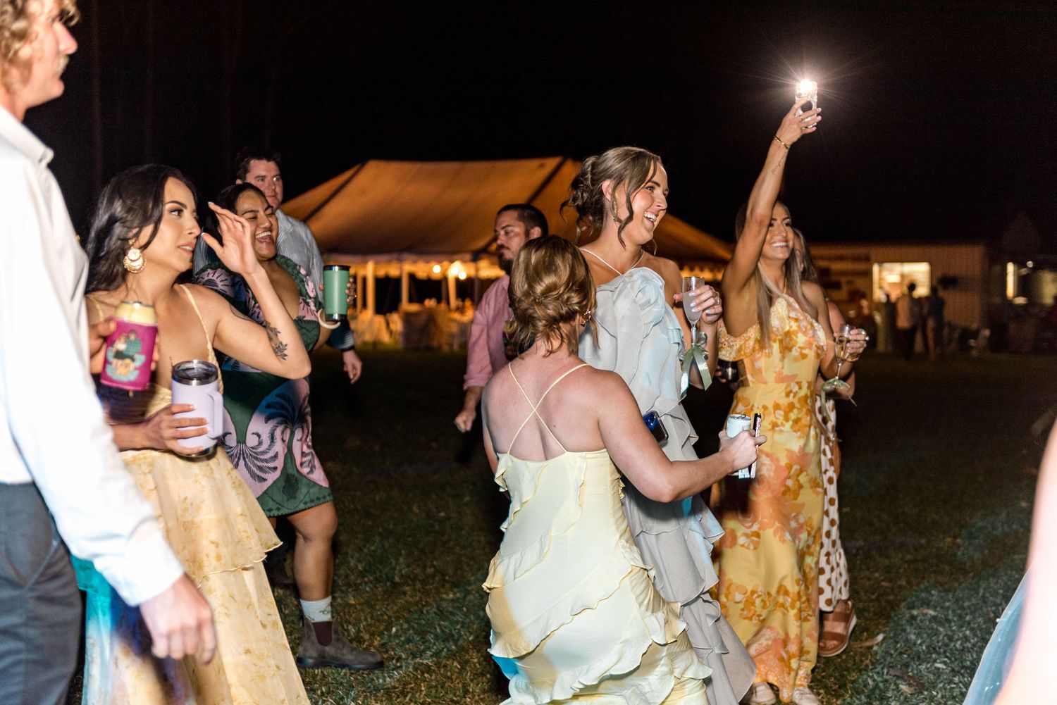 Bride raising sparkler while dancing with guests at outdoor evening wedding reception.