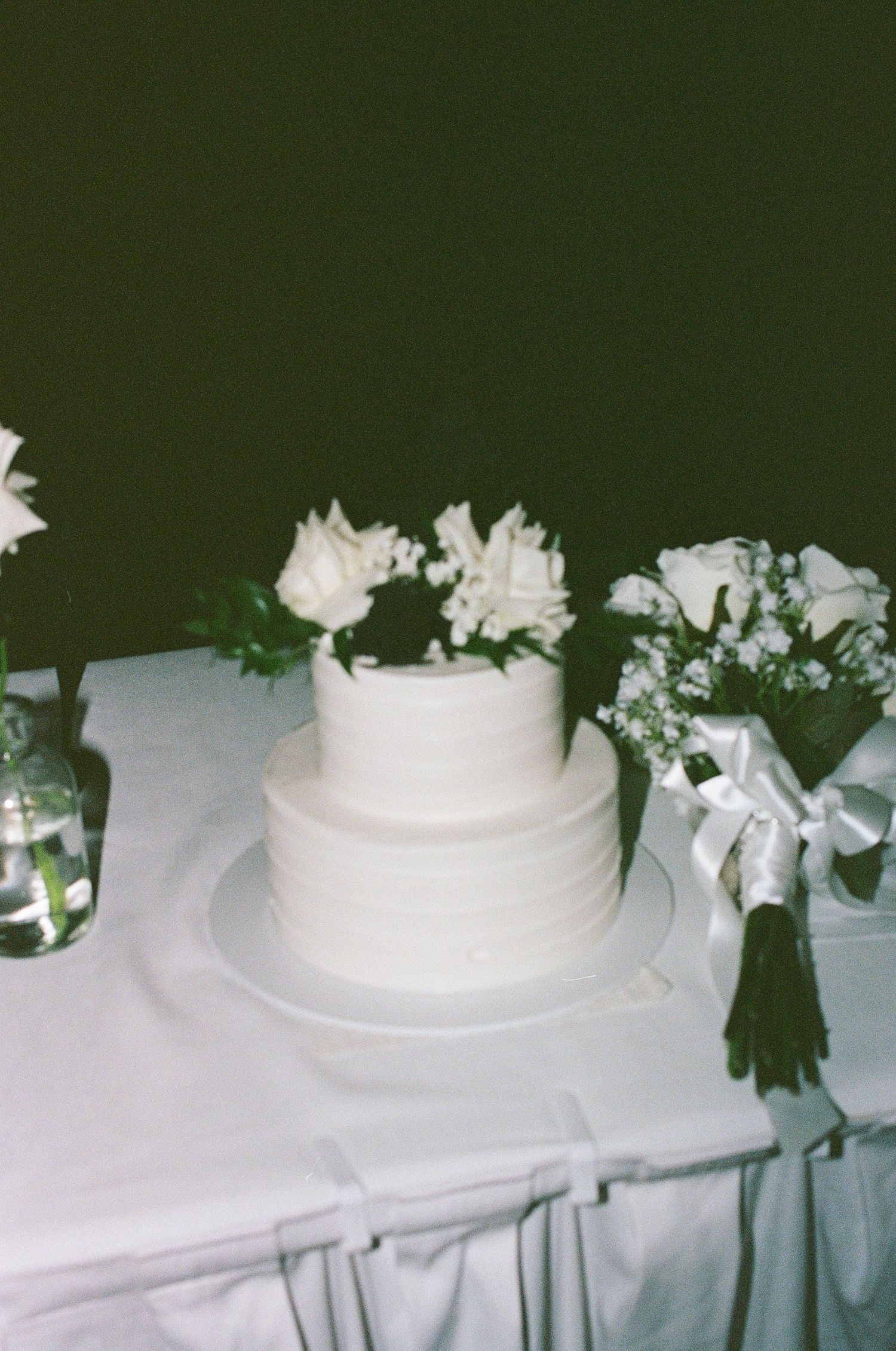 Elegant two-tier white wedding cake decorated with white roses on draped table.