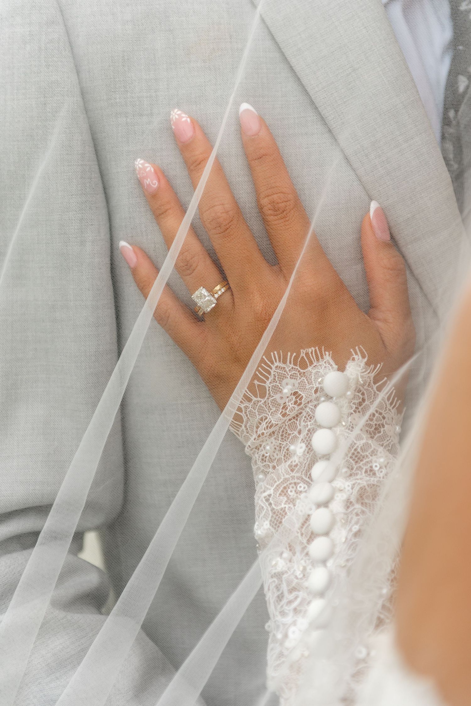 Close-up of bride's hand with diamond ring and pink French nails resting on groom's gray suit.