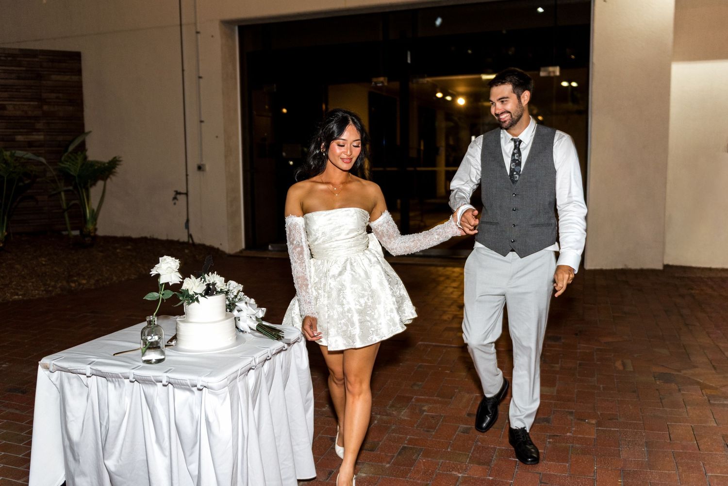 Bride in white mini dress and groom in gray suit walk hand in hand at reception.