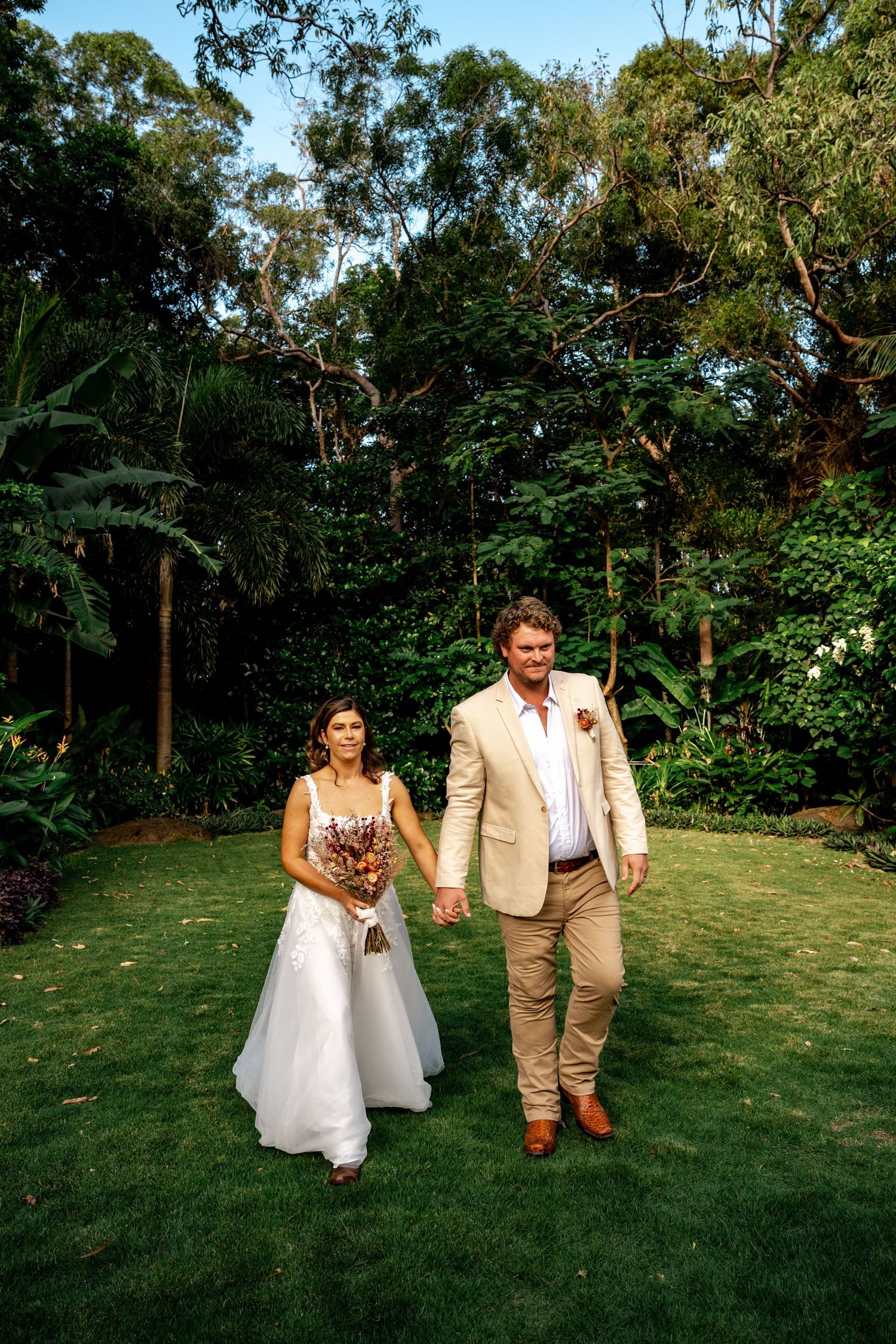 Newlywed couple walks hand in hand across green lawn with tropical garden background at dusk.