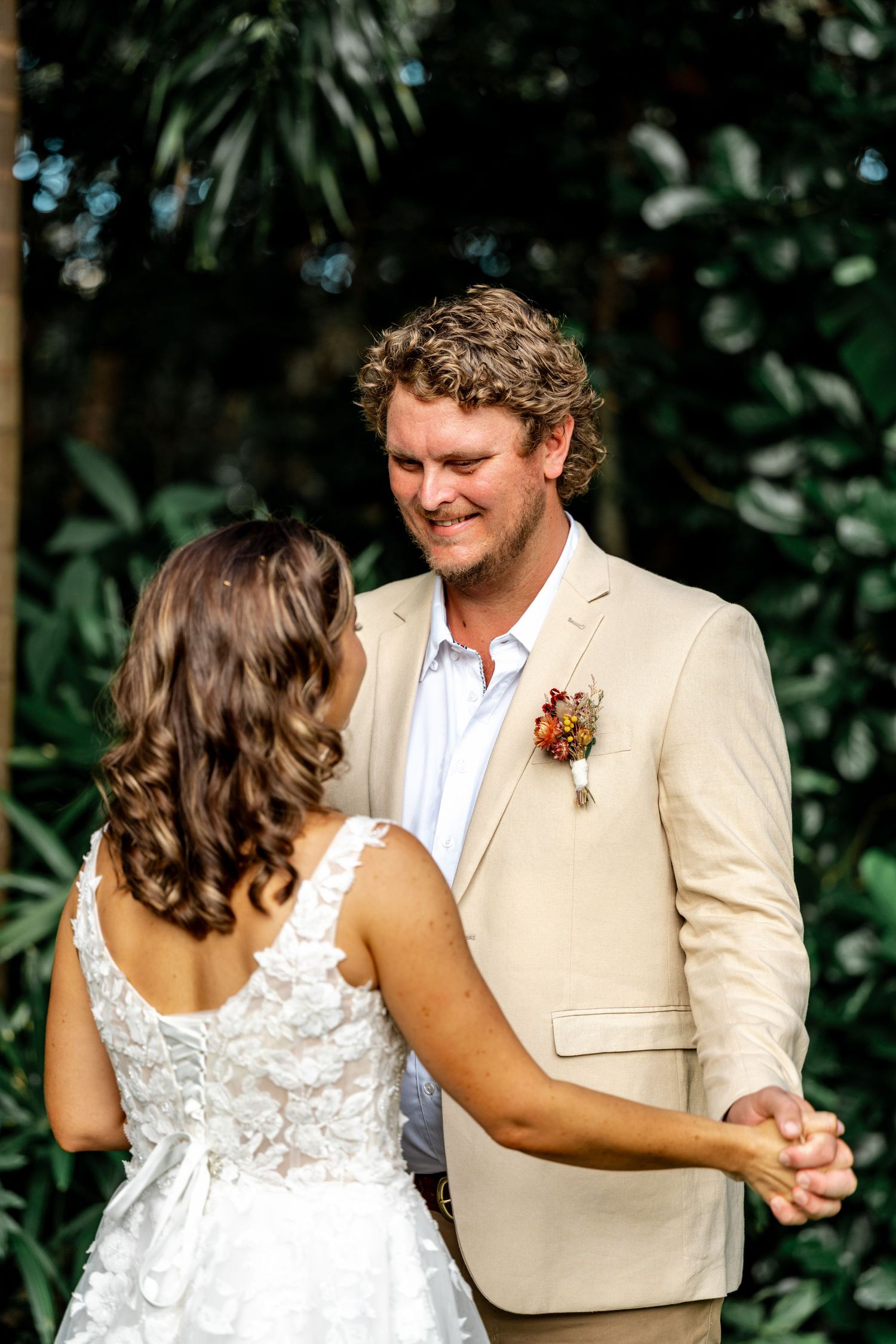 Groom in beige suit smiles at bride during outdoor wedding ceremony surrounded by greenery.