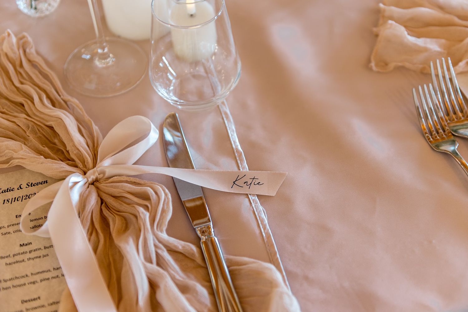 Wedding place setting with name card, satin ribbon napkin, silverware, and champagne glass on blush tablecloth.