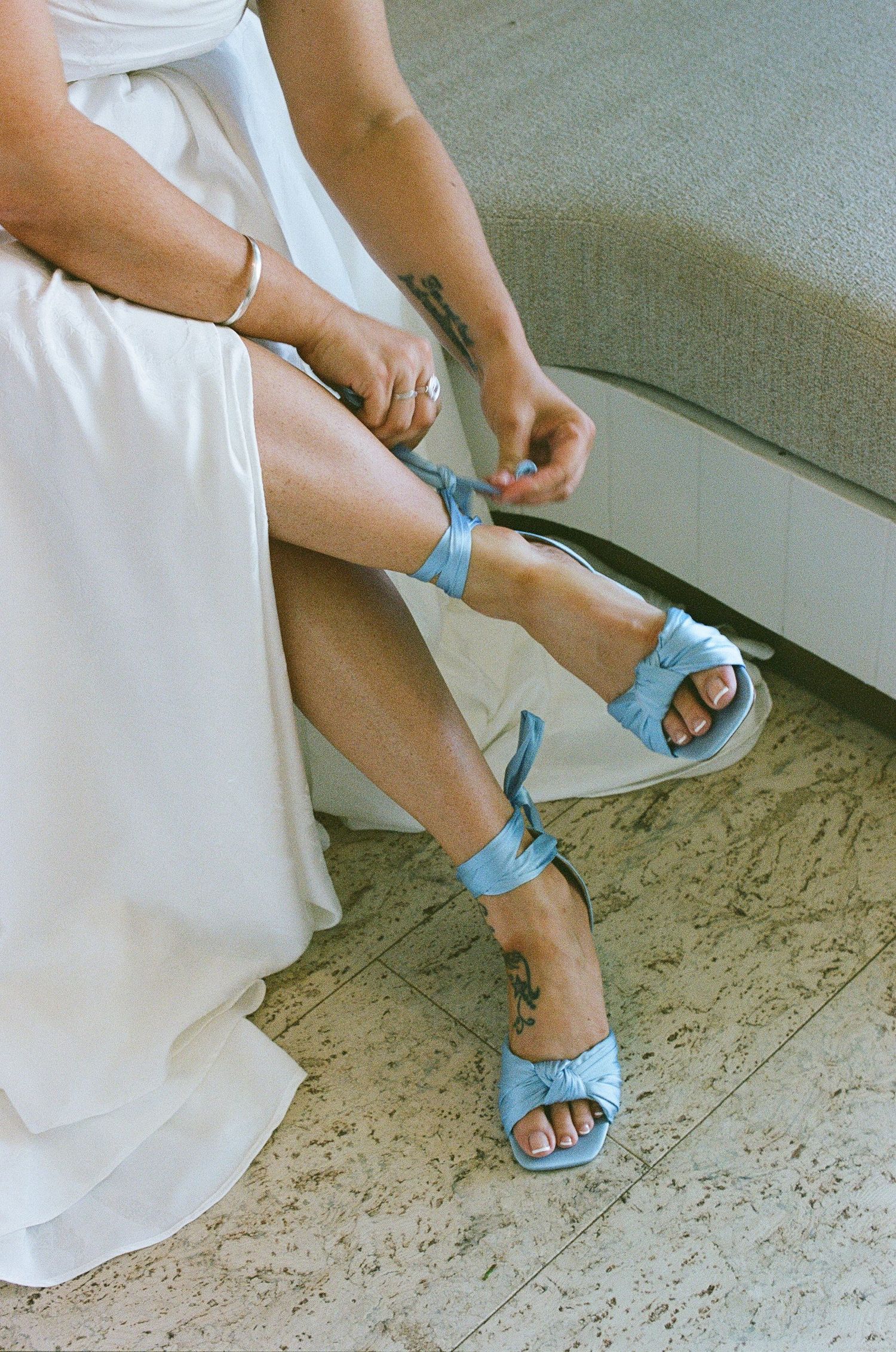 Close-up of bride fastening light blue satin heeled sandals with ankle ties on marble floor.