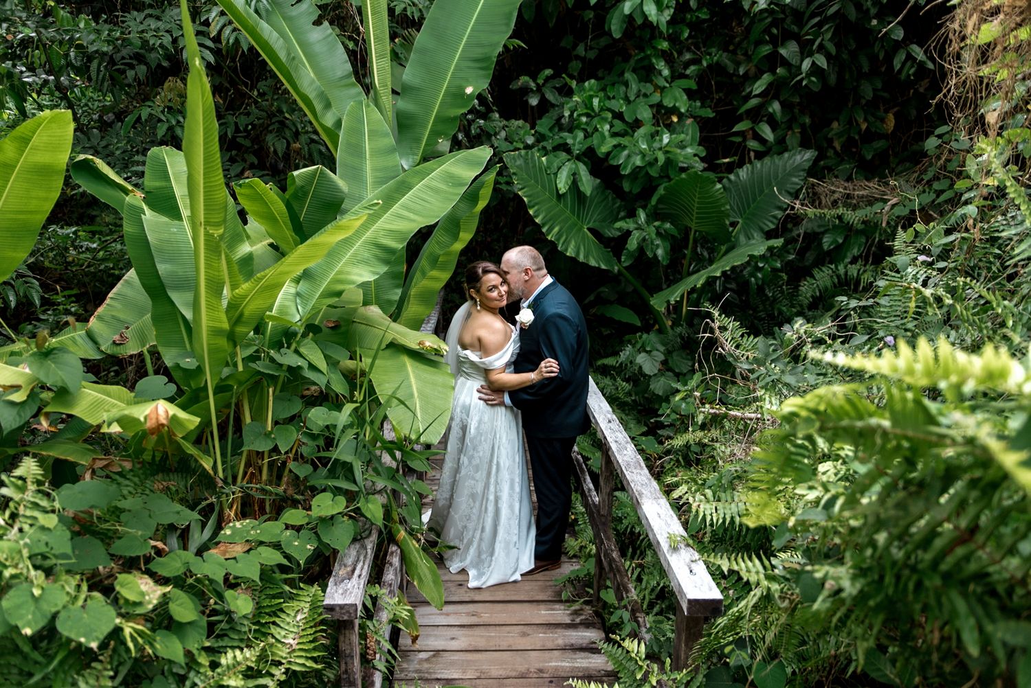 Bride and groom embracing on wooden bridge amid vibrant tropical foliage and banana trees.