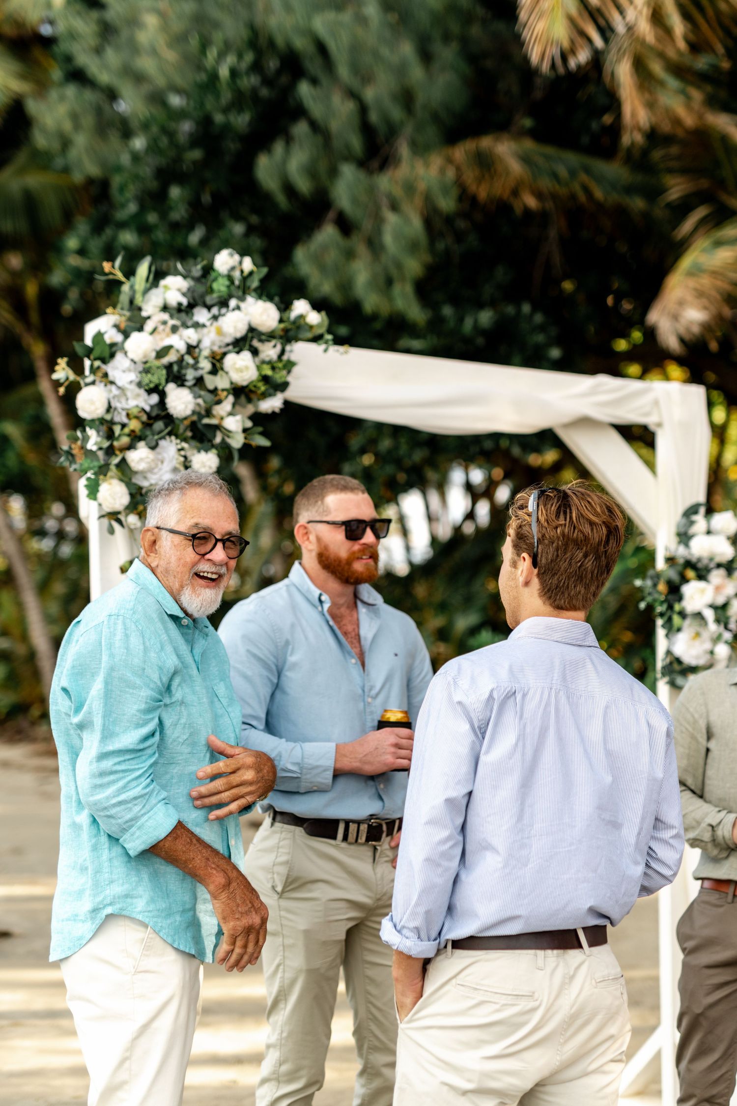 Three men in casual beach wedding attire chat and laugh near a floral arch with white blooms outdoors.