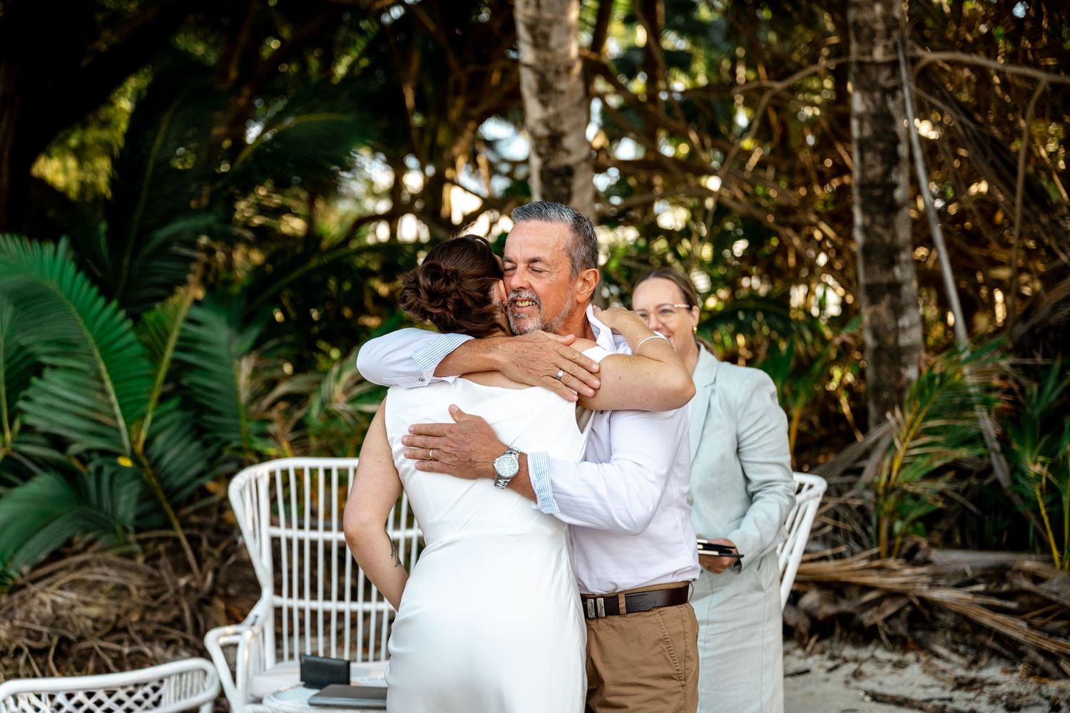 Bride in white gown hugging an older man on a sandy beach surrounded by tropical greenery at Four Mile Beach, Port Douglas.
