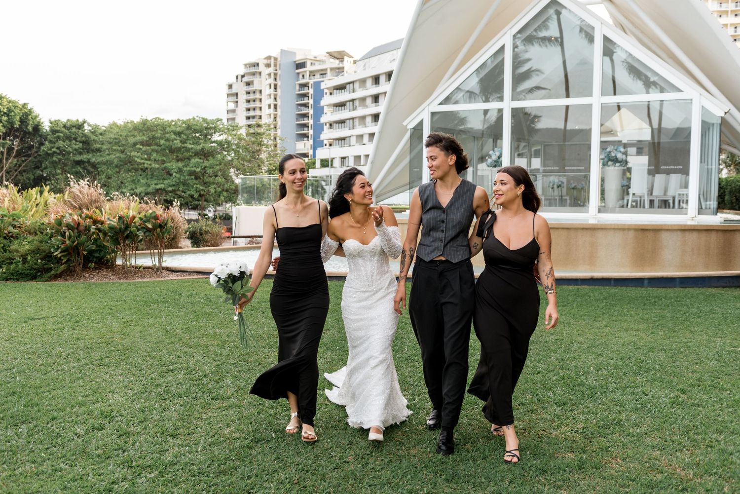Bride in white gown walking with bridesmaids in black dresses at outdoor wedding venue.