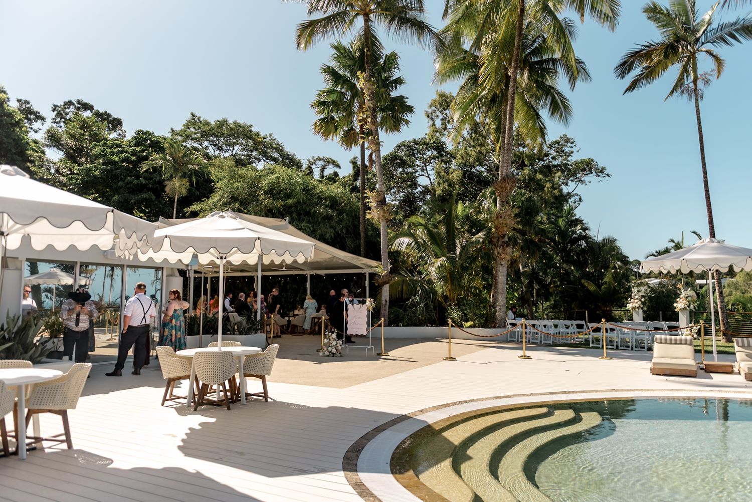 Guests mingling poolside at a tropical venue with white marquee umbrellas, palm trees, and bright blue skies.