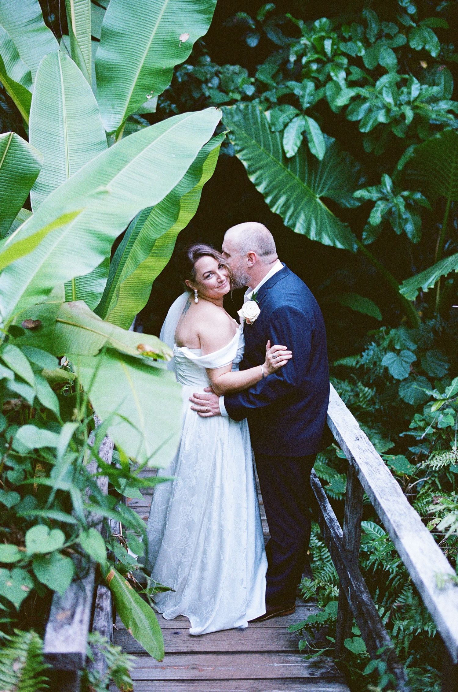 Bride and groom sharing intimate moment on wooden bridge surrounded by lush tropical banana and fern plants.