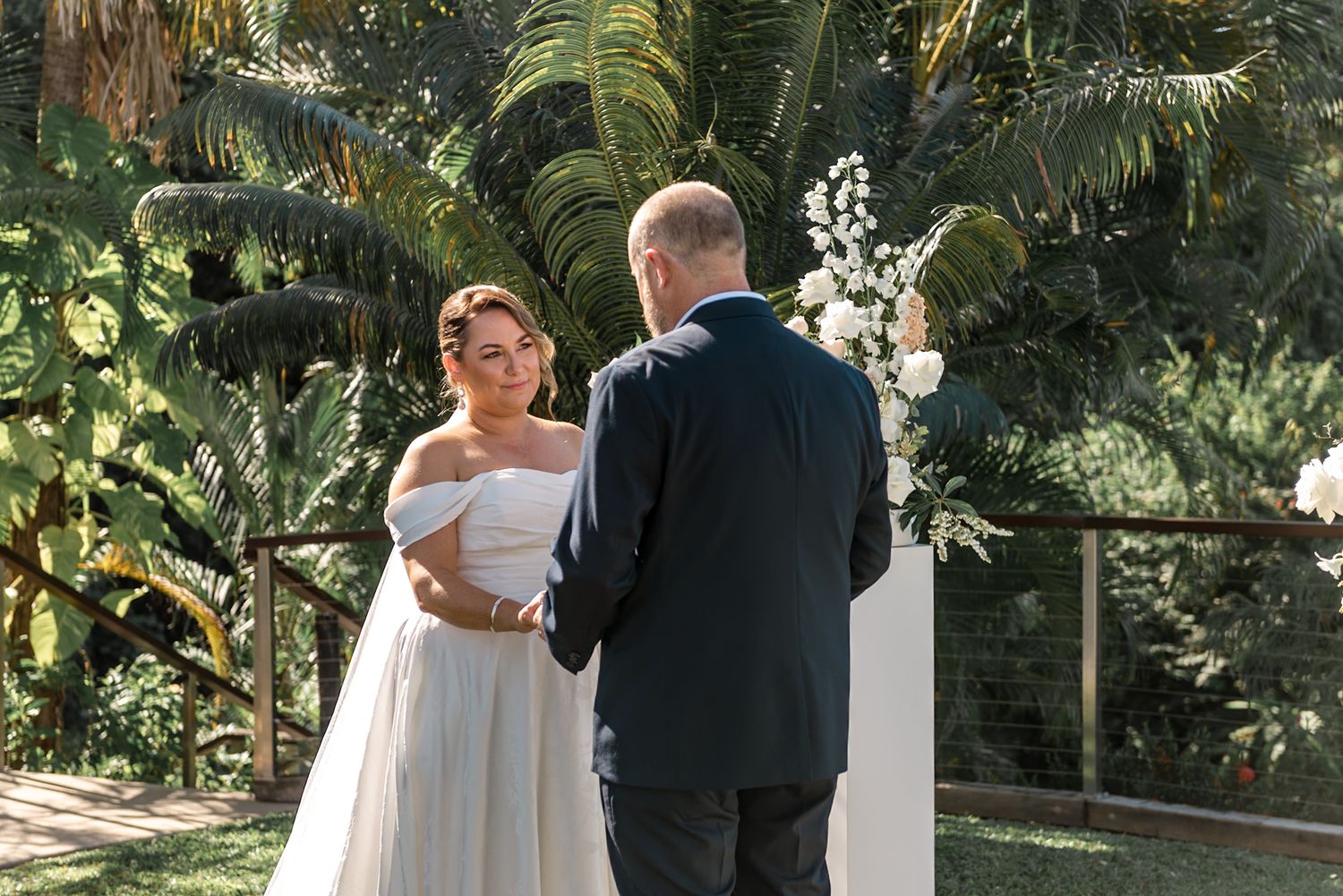Couple exchanges vows at sunlit outdoor wedding ceremony with lush tropical palm trees in the background.