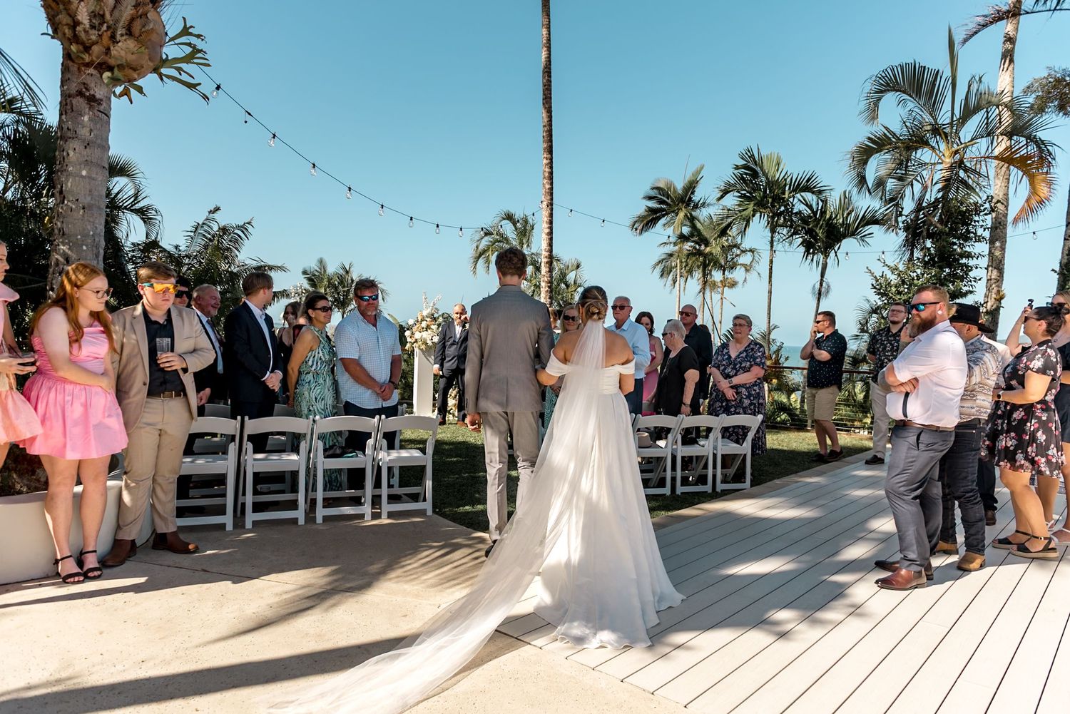 Bride and groom stand at outdoor wedding altar surrounded by guests seated in white chairs under palm trees.