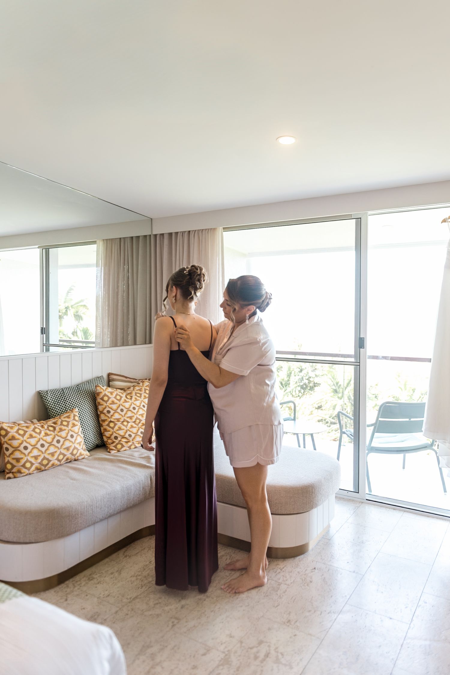Woman in burgundy gown being helped by another in white robe in a bright modern hotel room.
