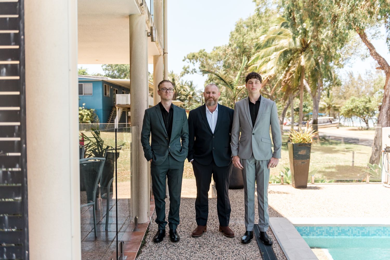 Three men in suits posing together outdoors near a pool with palm trees in the background.