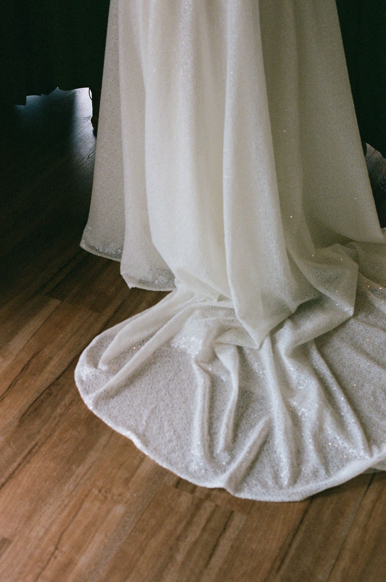 Close-up of a white sparkly wedding dress train spread elegantly on a hardwood floor.