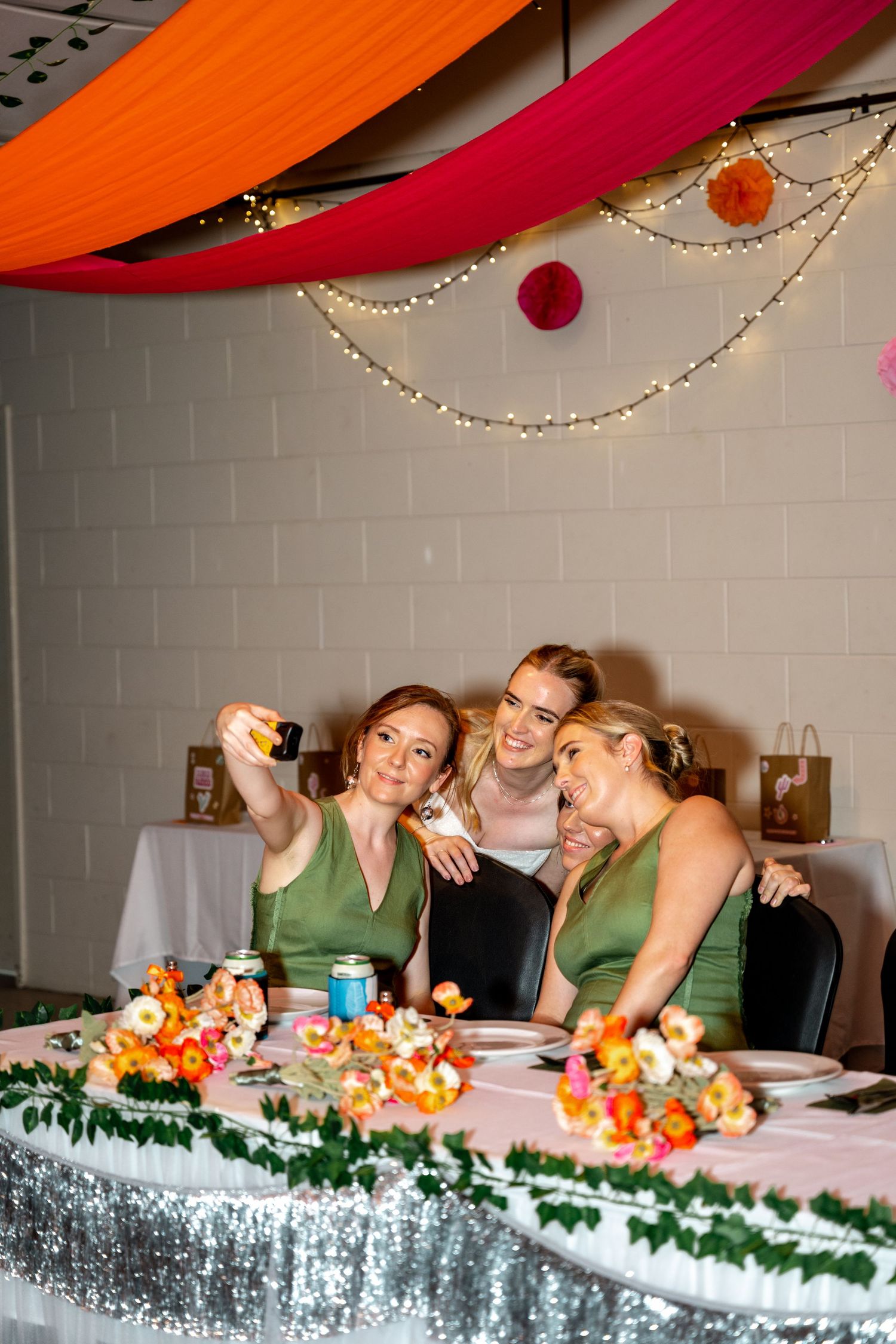 Three bridesmaids in green dresses taking a selfie at the decorated wedding reception head table.