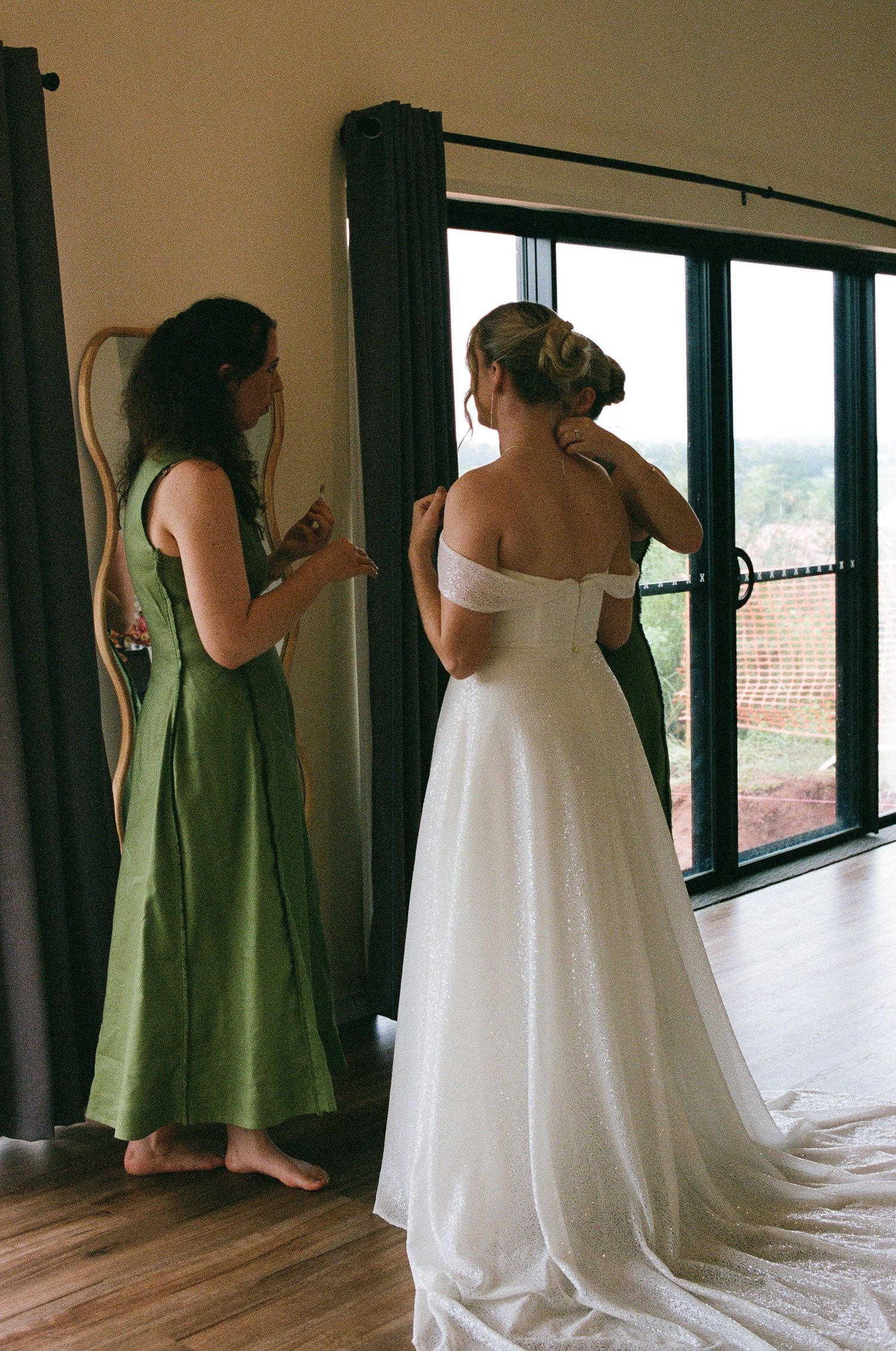 Bridesmaid in green linen dress helps bride with off-shoulder white wedding gown near window.