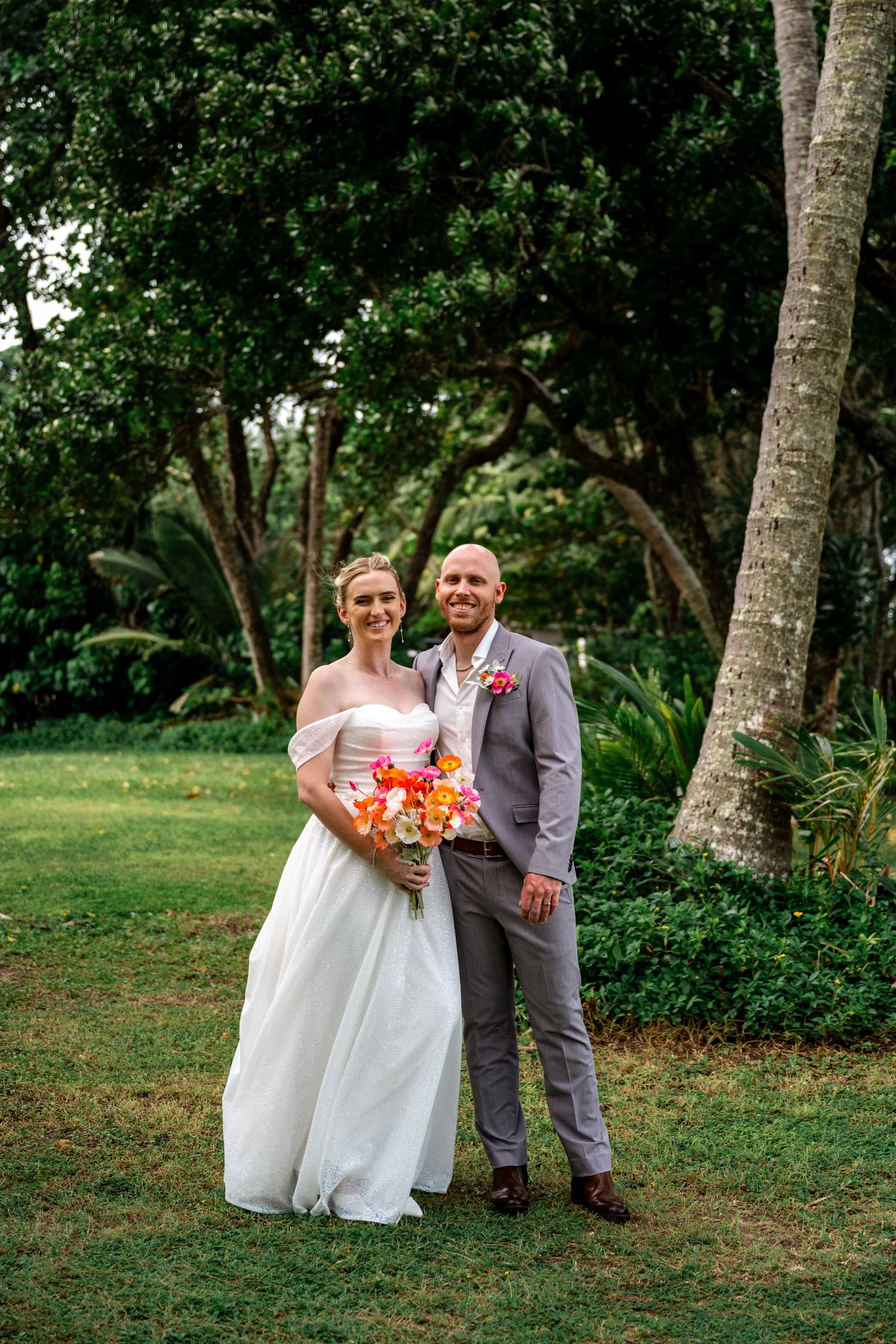 Bride and groom smiling together in lush tropical garden, bride holding vibrant orange bouquet in white off-shoulder gown.