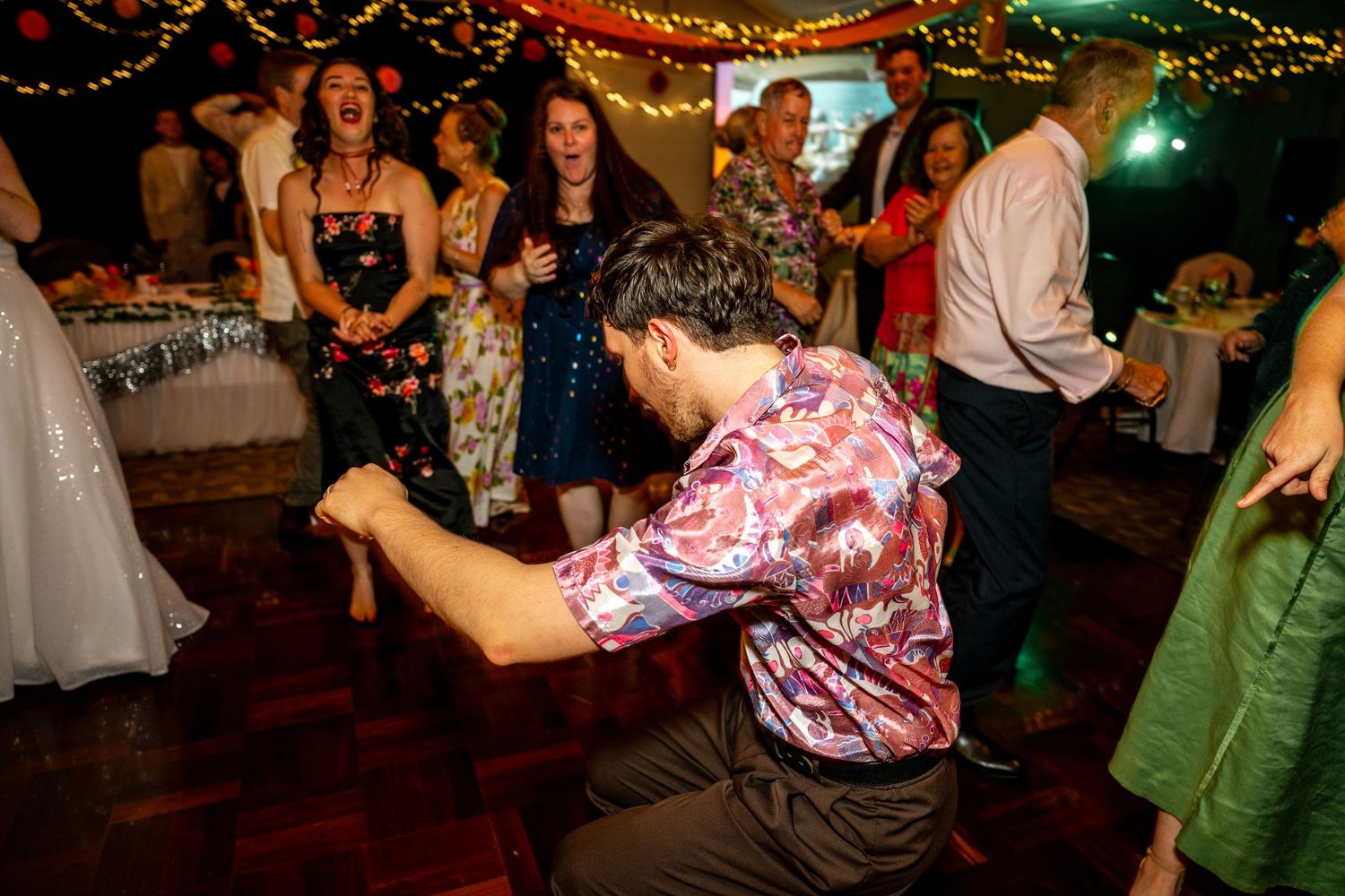 Wedding guest in floral shirt dances enthusiastically on crowded dance floor at festive reception.