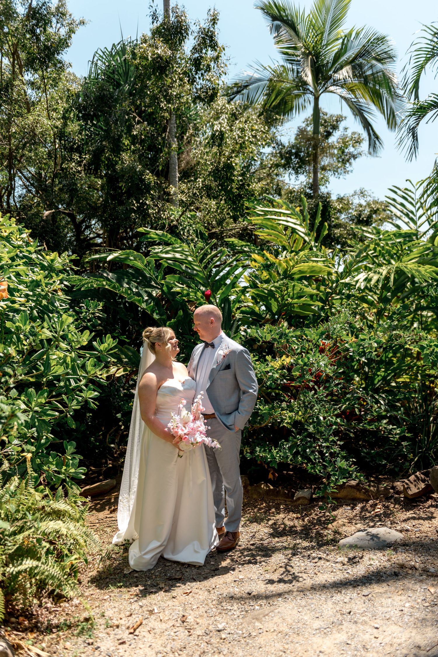 Bride and groom stand together on a garden path surrounded by vibrant tropical plants and tall palm trees.